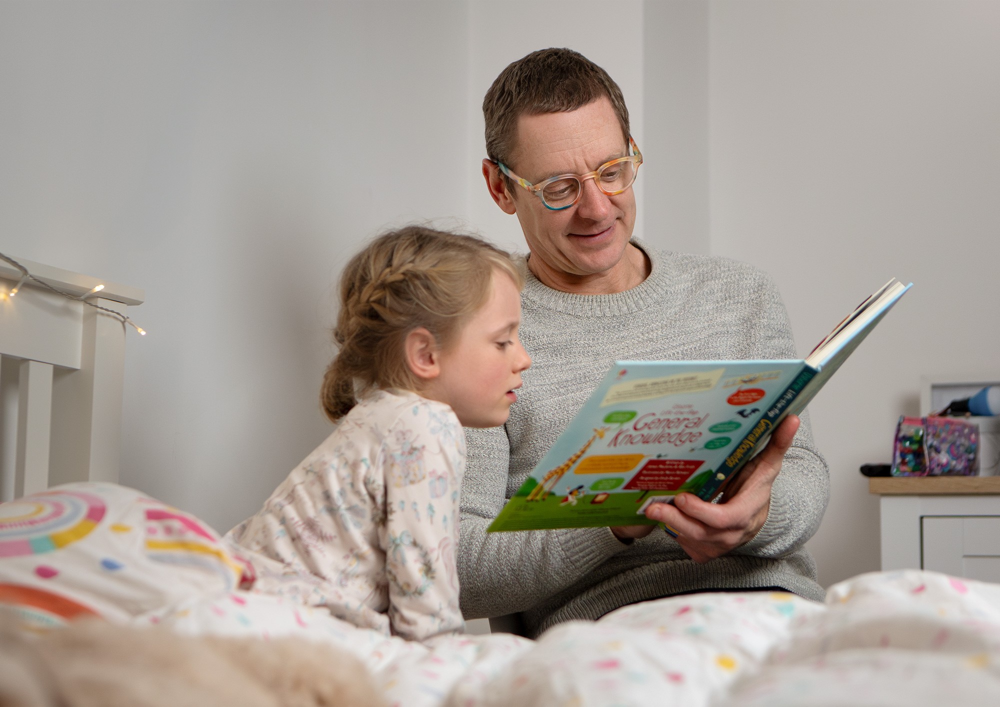 A father wearing colourful speckeled Izipizi glasses reading his daughter a bedtime story, photographed by Matthew Brown.