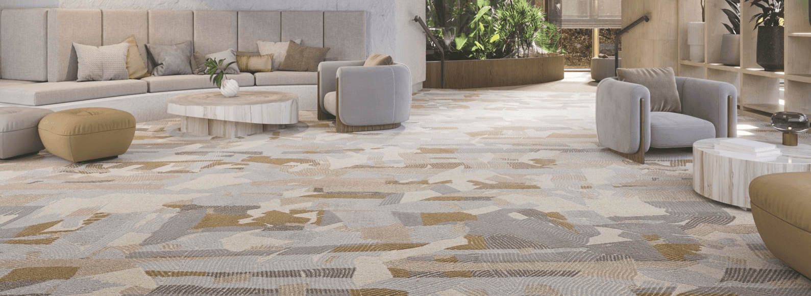 A bright, airy hotel lobby with a large-scale patterned carpet in warm gold, taupe, and soft grey tones. The space features organic modern furnishings, wood-paneled ceilings, and floor-to-ceiling windows with lush greenery beyond.