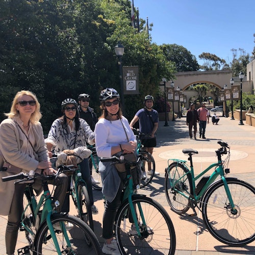 一群头戴头盔、骑自行车的人站在一个阳光明媚、户外广场的拱形入口附近，背景是一片树木。