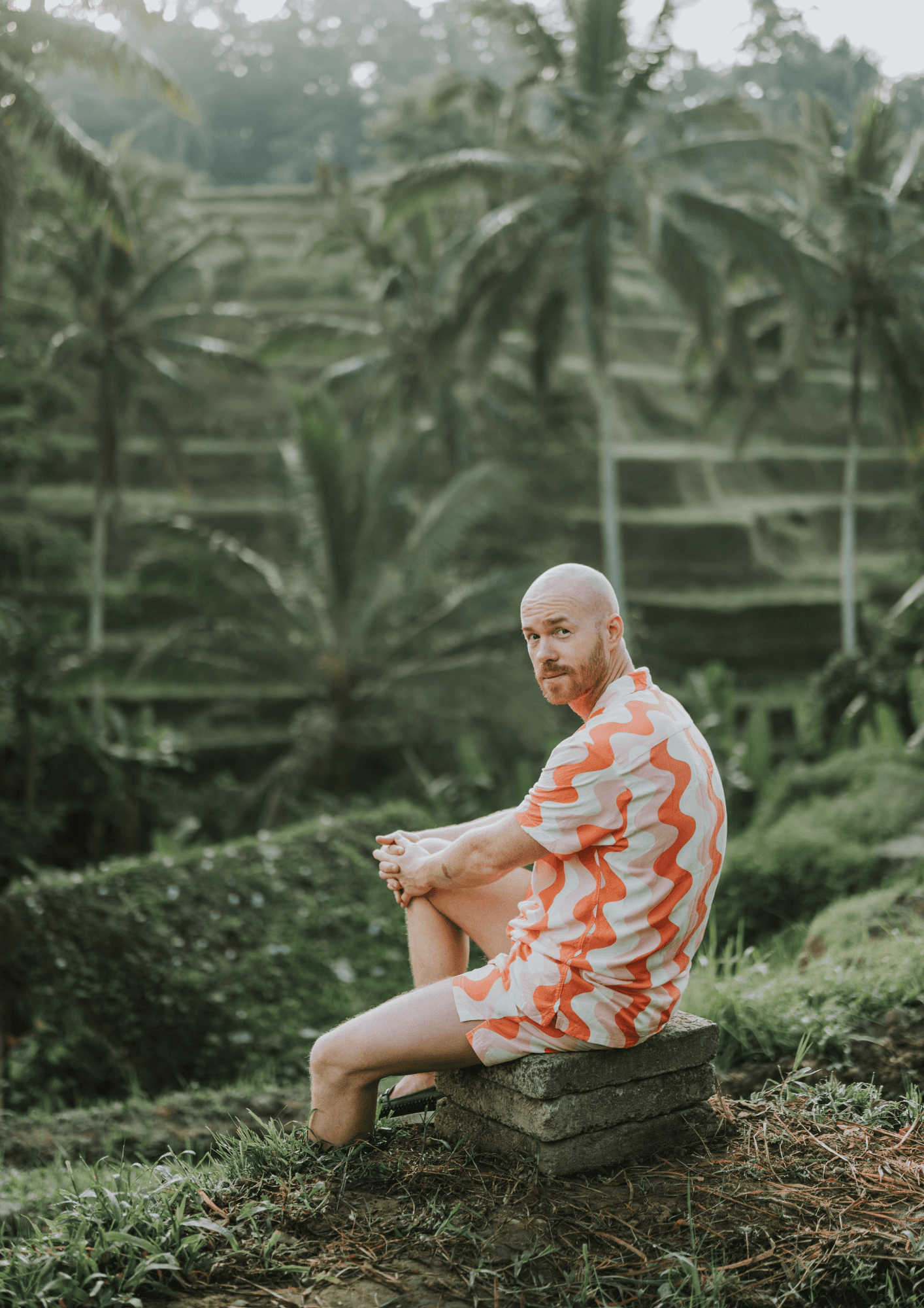 Thom Rikko seated outdoors amid palm trees and terraced greenery.