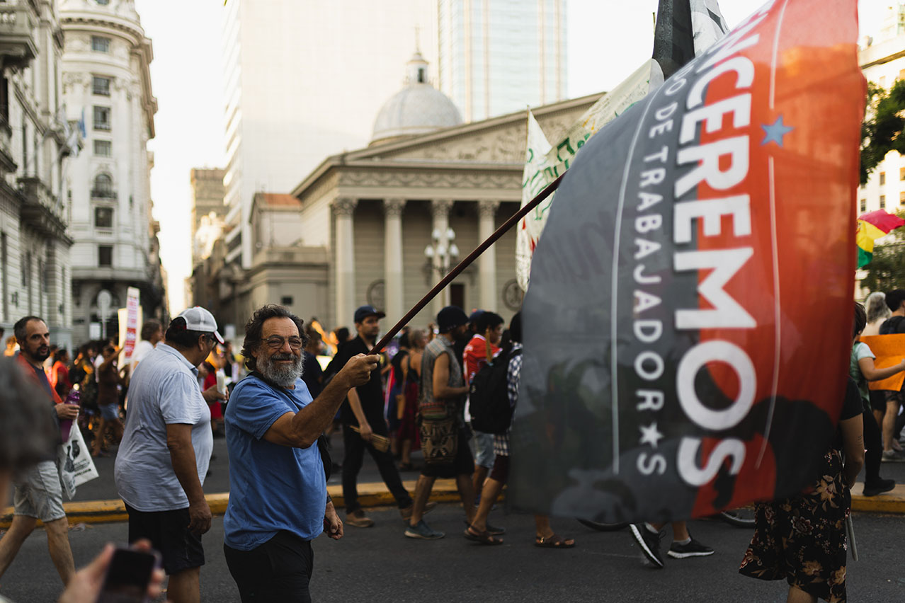 Old man holding a flag during a manifestation in Buenos Aires, Argentina, with sunlight hitting building tops.