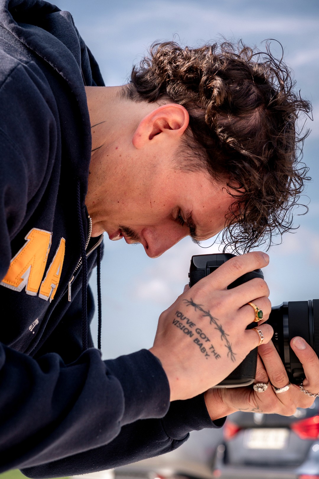 Plan serré sur un photographe, tatouage visible, ciel bleu en fond.