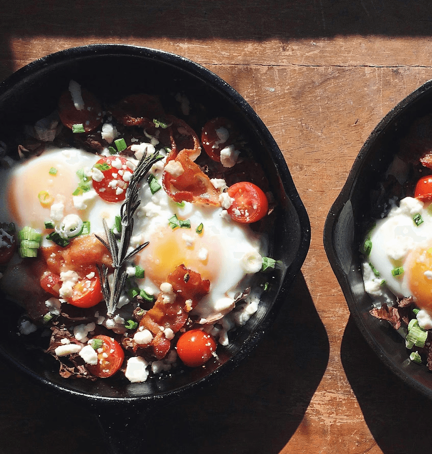Deux poêles en fonte avec des œufs au plat, tomates, feta, bacon, oignons verts et romarin sur une table en bois ensoleillée.