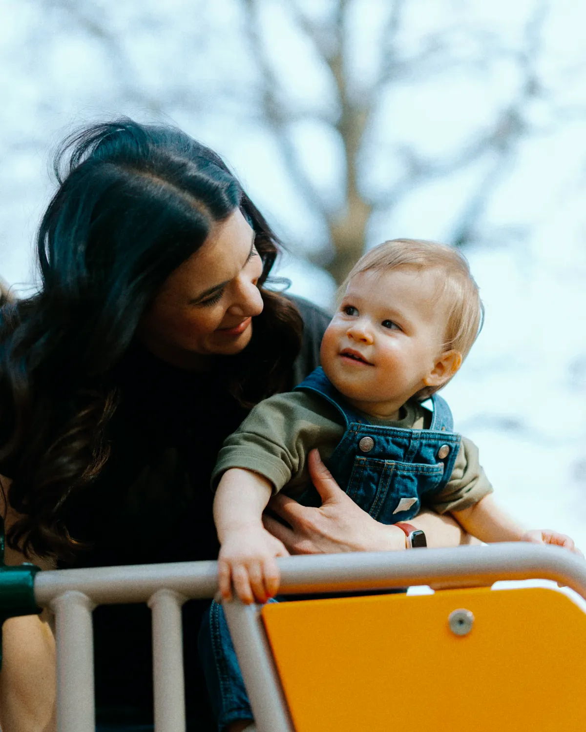Mom with dark hair on a playground holding her son while looking at him