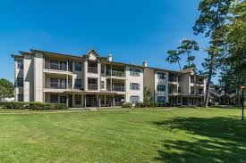 A large, three-story apartment or condominium building with light beige siding and numerous balconies, viewed from a wide, well-maintained green lawn. The building is situated under a clear blue sky, with tall pine trees visible on the right side.