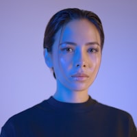 Portrait of a person with dark hair pulled back, wearing a black crewneck shirt, illuminated by blue and purple gradient lighting against a softly lit background.