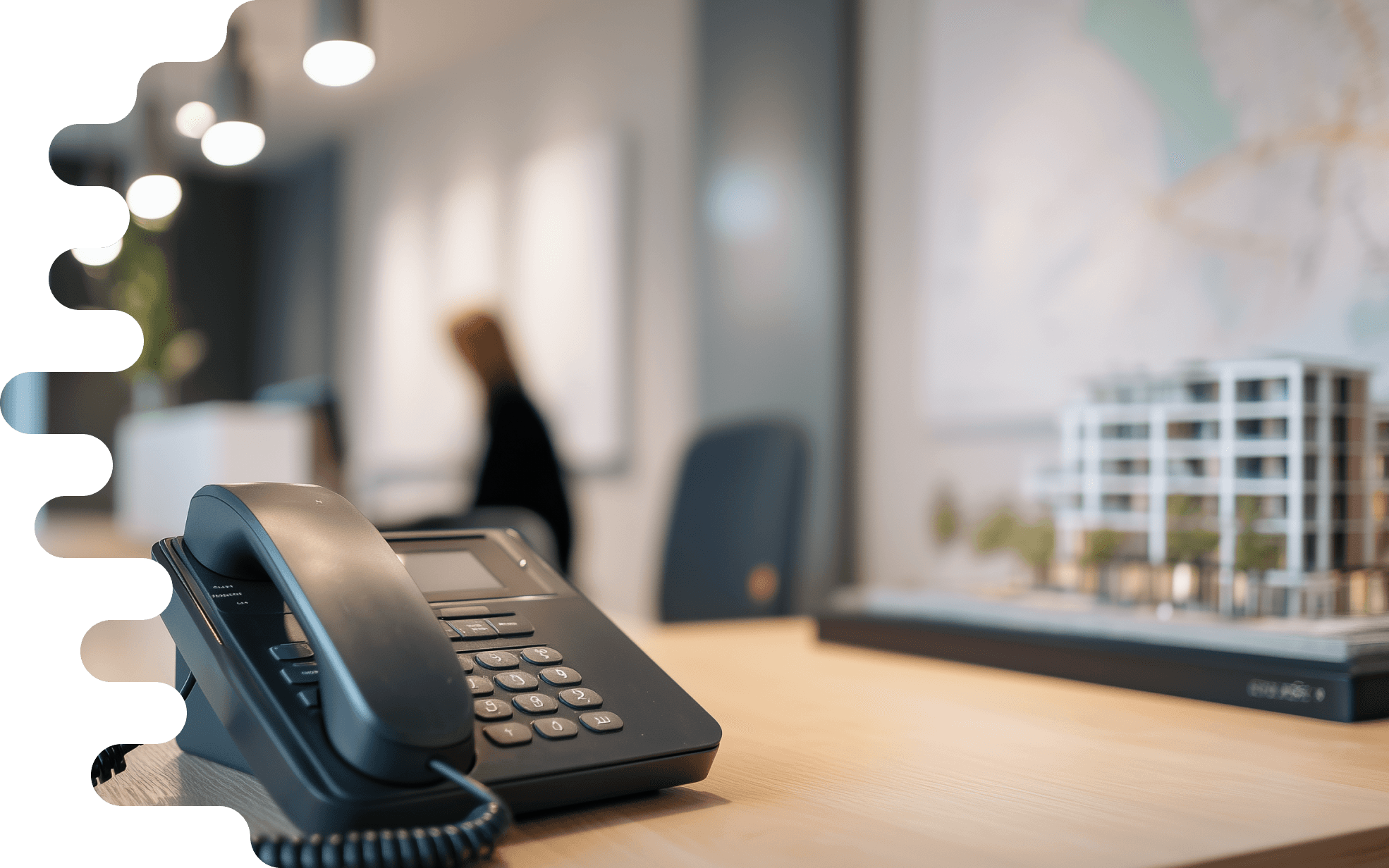"The image shows a modern office interior with a light wood desk. In the foreground, there is a black phone lying on the table. It has a large keypad and belongs to a typical office phone. In the background, a blurred person can be seen, who is apparently standing at a reception desk or busy working in the office. The desk is neat and tidy. To the right of the phone is an architectural model of a multi-story building, displayed on a dark base. The model features many windows and a green environment depicted with small trees. In the background, a wall with geometric maps or plans in soft colors can be seen. Above the table, some modern light sources hang, creating a bright and inviting atmosphere."