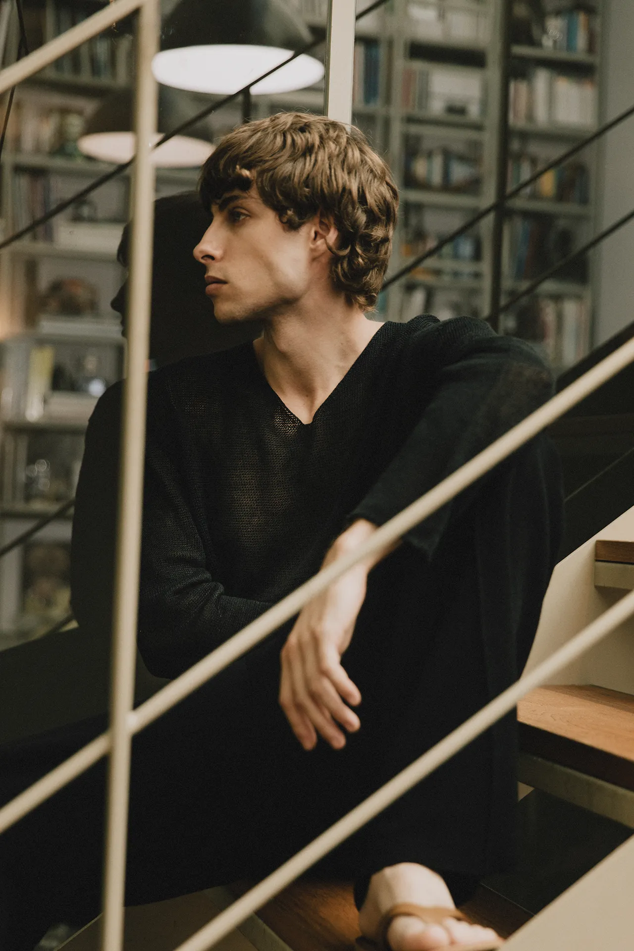 Annapurna SS26 collection men's black sheer V-neck sweater worn by a model sitting on stairs.