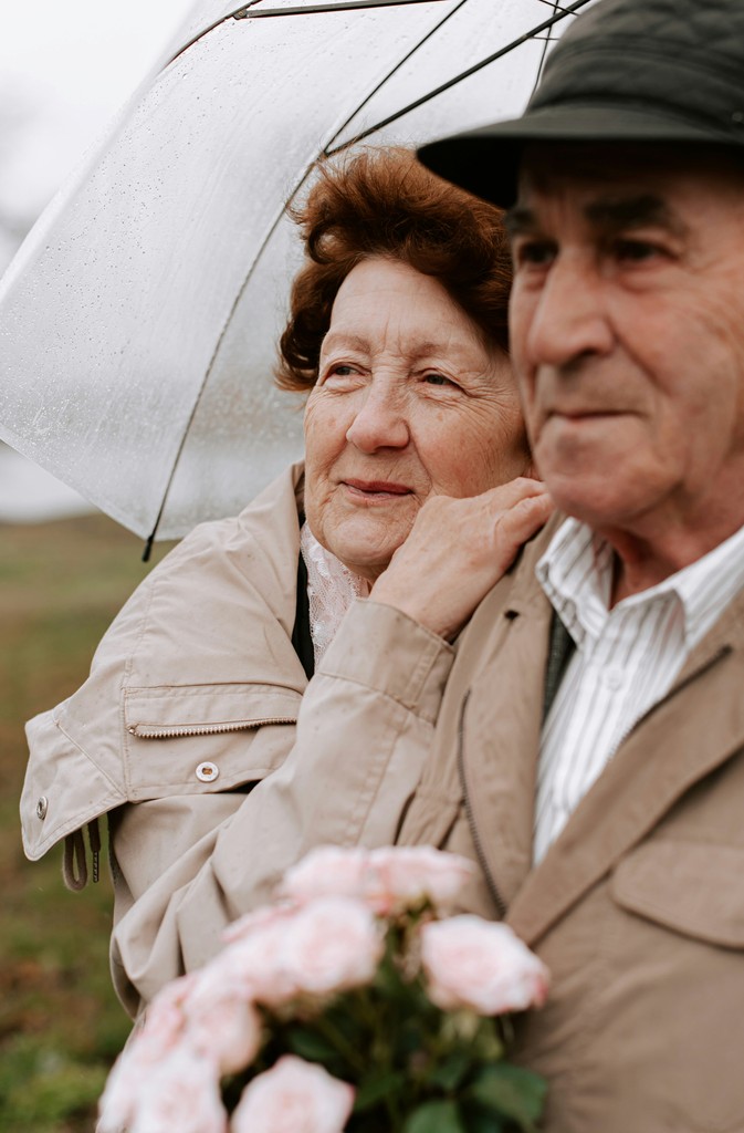 An elderly couple stands close together under an umbrella on a rainy day, with the woman gently holding her partner and a bouquet of pink roses, symbolizing enduring love and companionship.