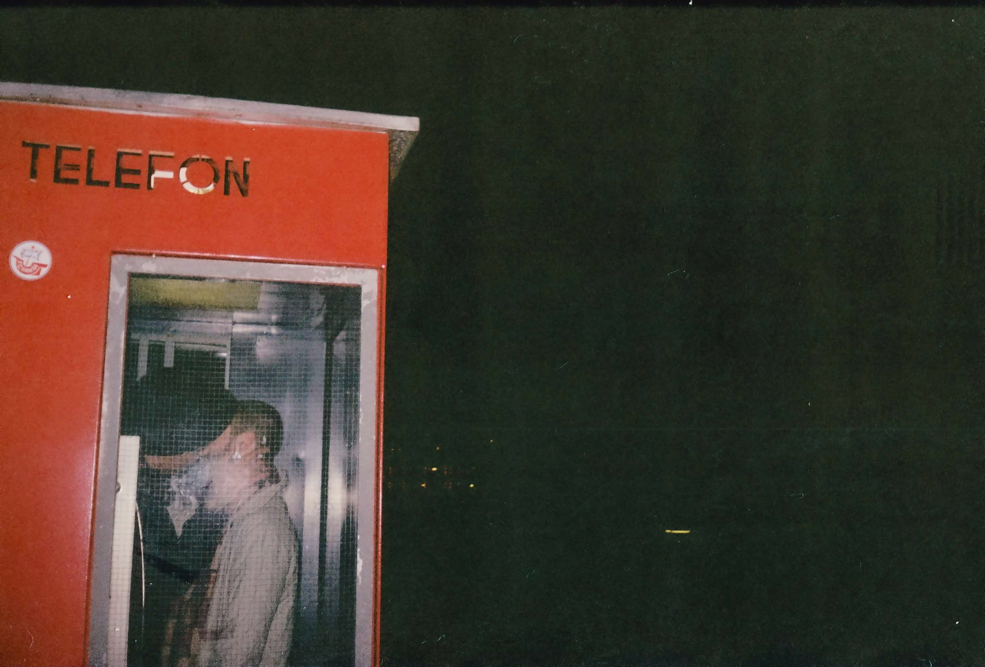 A person stands inside a vintage red telephone booth at night, glowing softly under artificial light against a dark background.