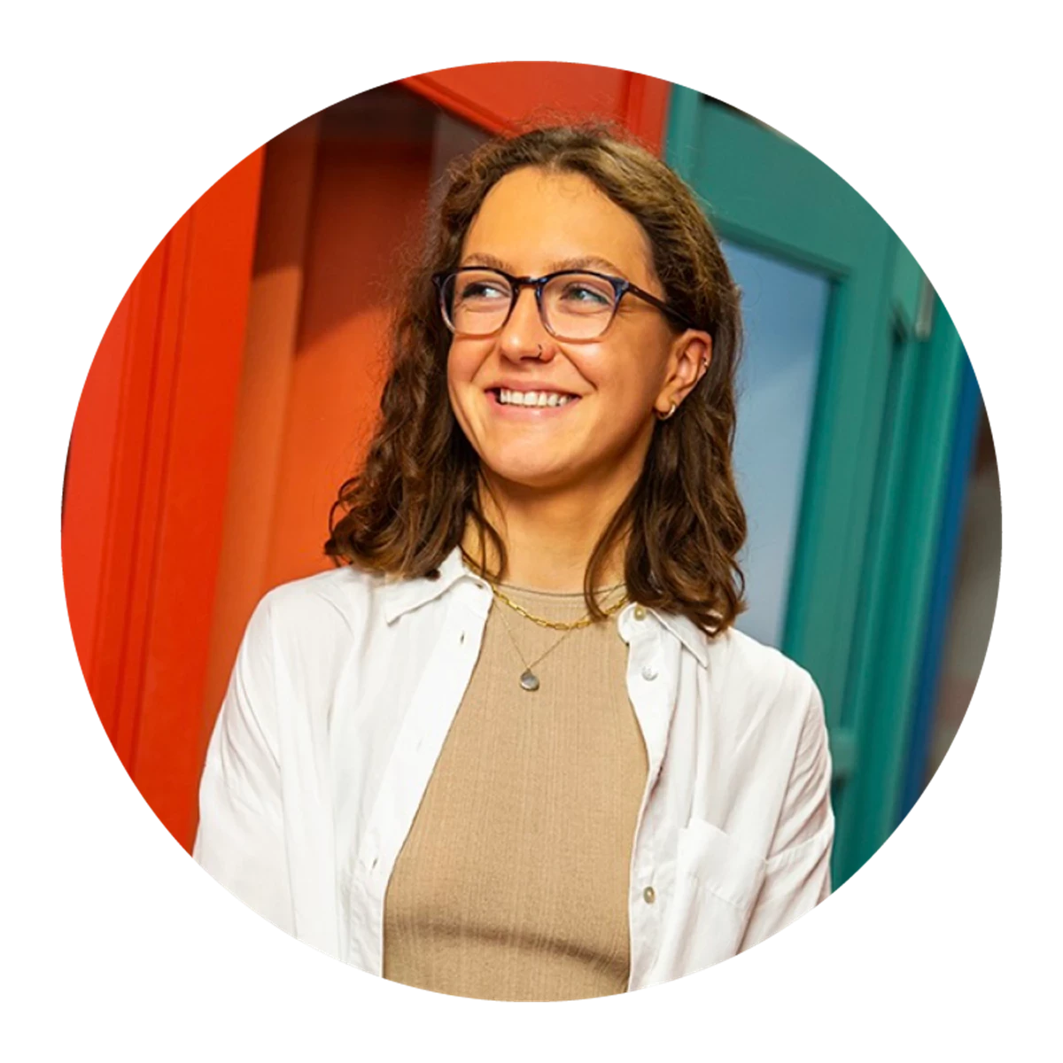 A person with curly hair wears a beige top layered with a white shirt. They stand in front of brightly coloured red and teal structures.