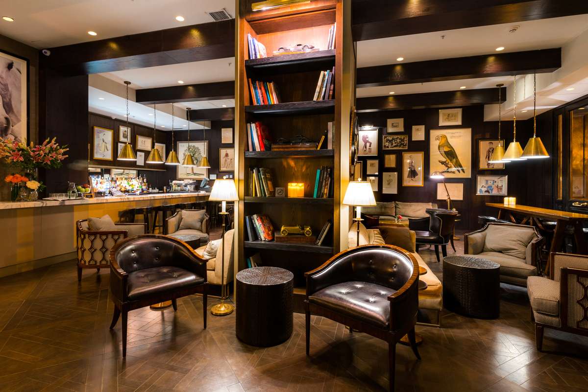 A cozy hotel lobby features leather armchairs, a tall wooden bookshelf, and warm lighting over a reception desk.