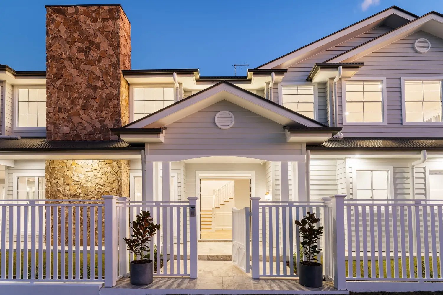 A modern two-story house with light siding, a prominent stone chimney, and a white picket fence at dusk.
