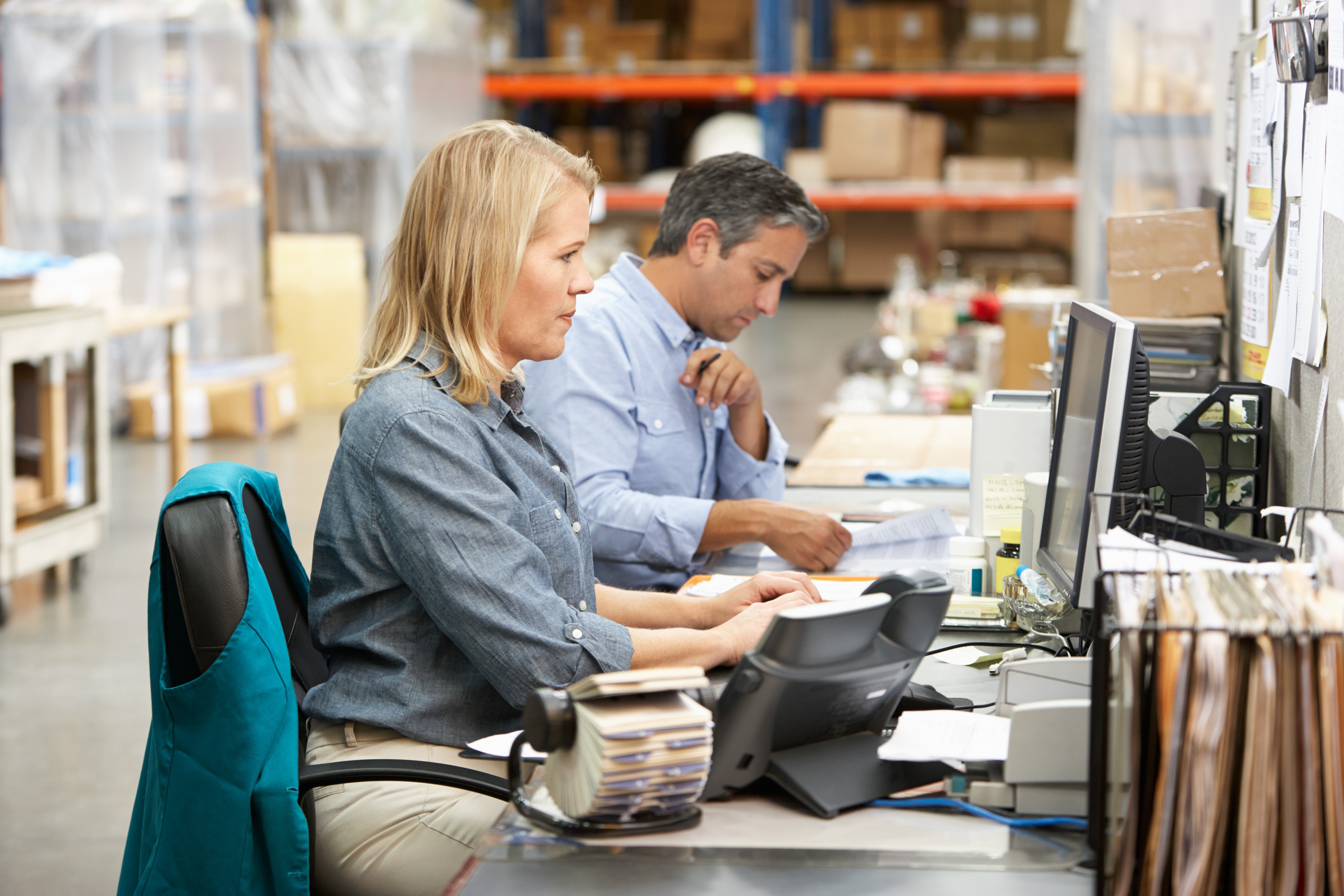 Warehouse office staff reviewing paperwork and entering data at computer workstations inside a 3PL facility