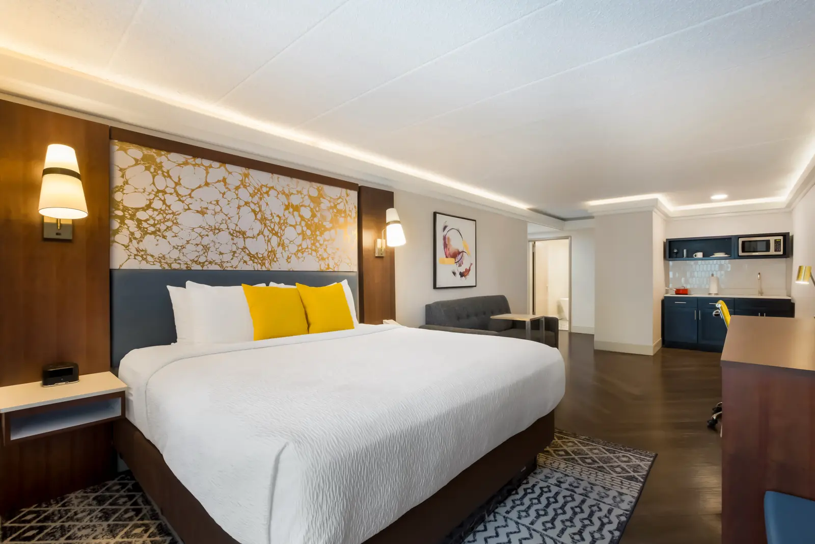 Modern hotel room with a large bed, desk, and kitchenette.
