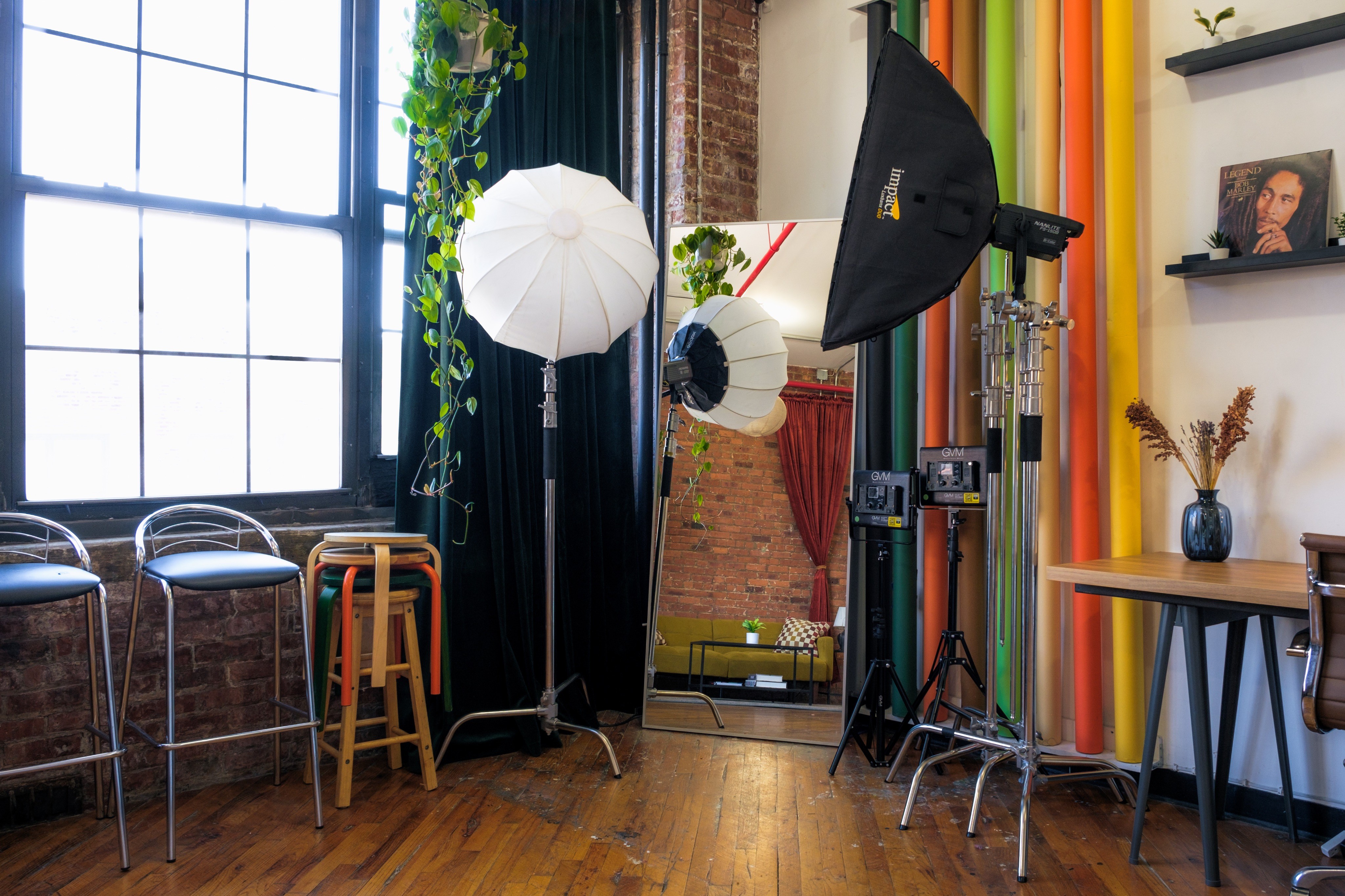 A daylight photography studio featuring a large wall of colorful backdrops, professional lighting equipment, and a workspace with shelves displaying decor and camera gear. A floral wall installation and a large mirror add artistic elements to the studio’s bright, versatile environment.