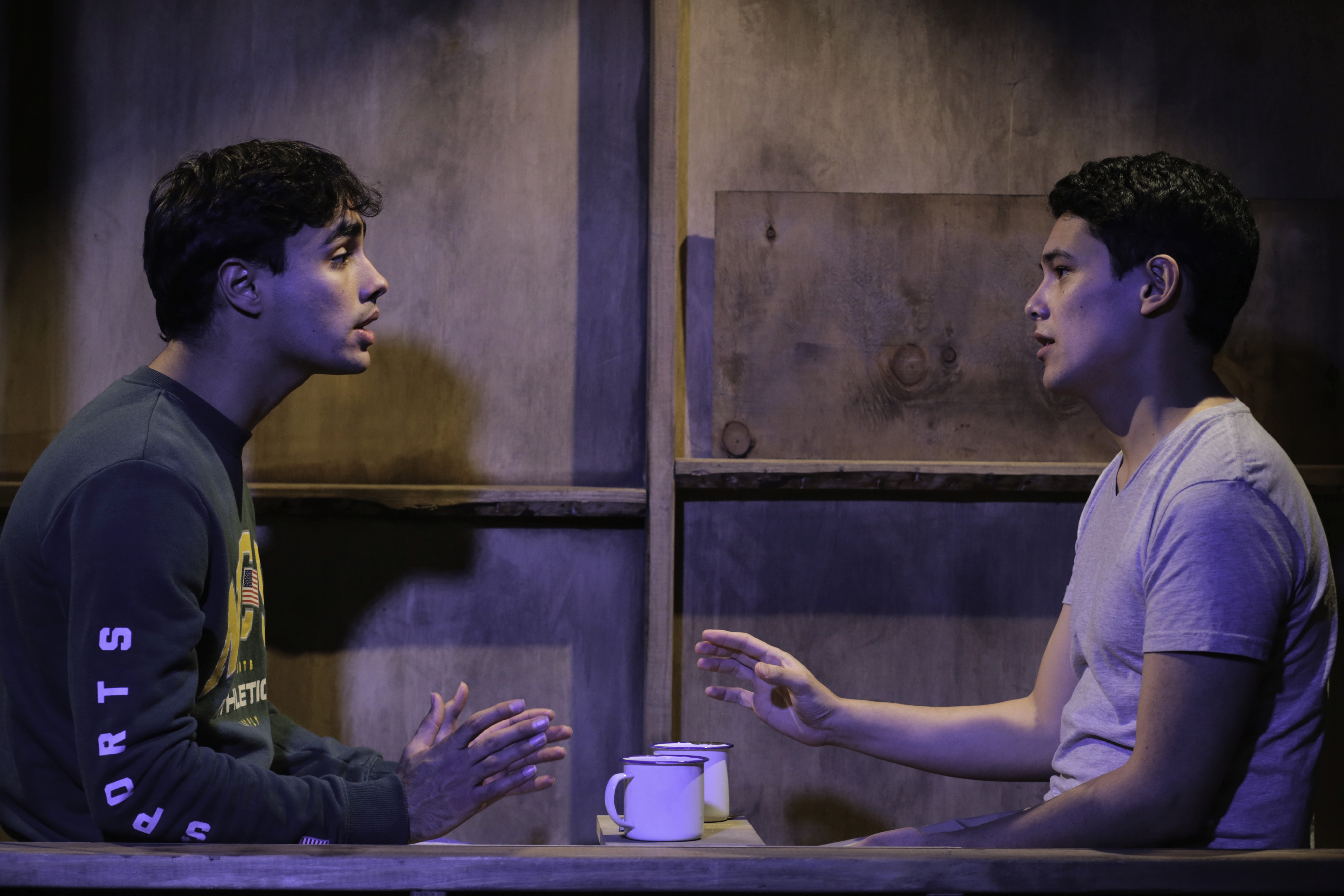 Two young men sit across from each other at a simple wooden table in a dimly lit setting, each with a white enamel cup in front of them. They appear to be in deep conversation, gesturing with their hands. One wears a green sweatshirt, the other a grey T-shirt, with rustic wooden walls behind them suggesting a sparse, intimate environment.