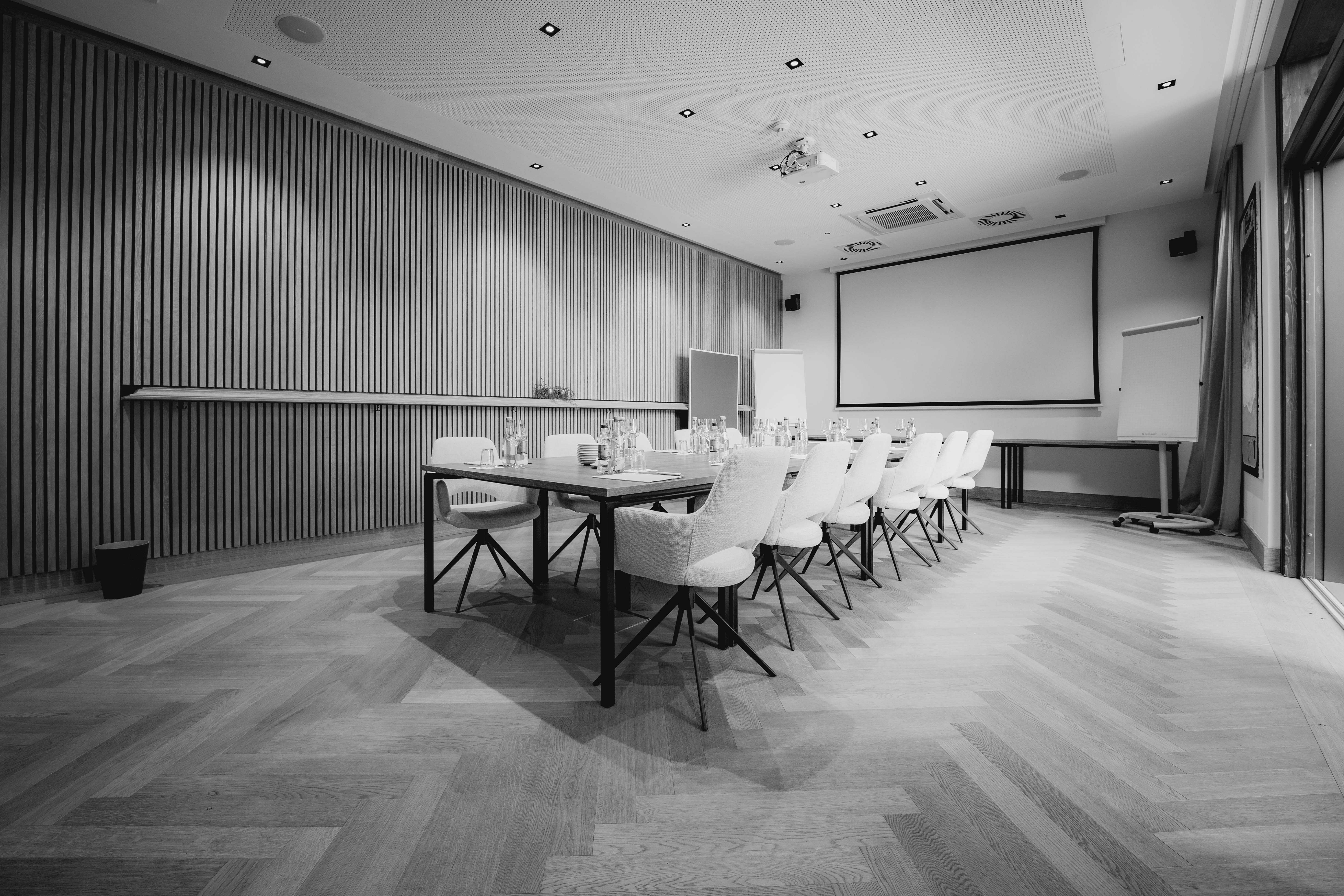 A modern conference room with a large screen, chairs, and tables set up for a meeting. Black and white image.