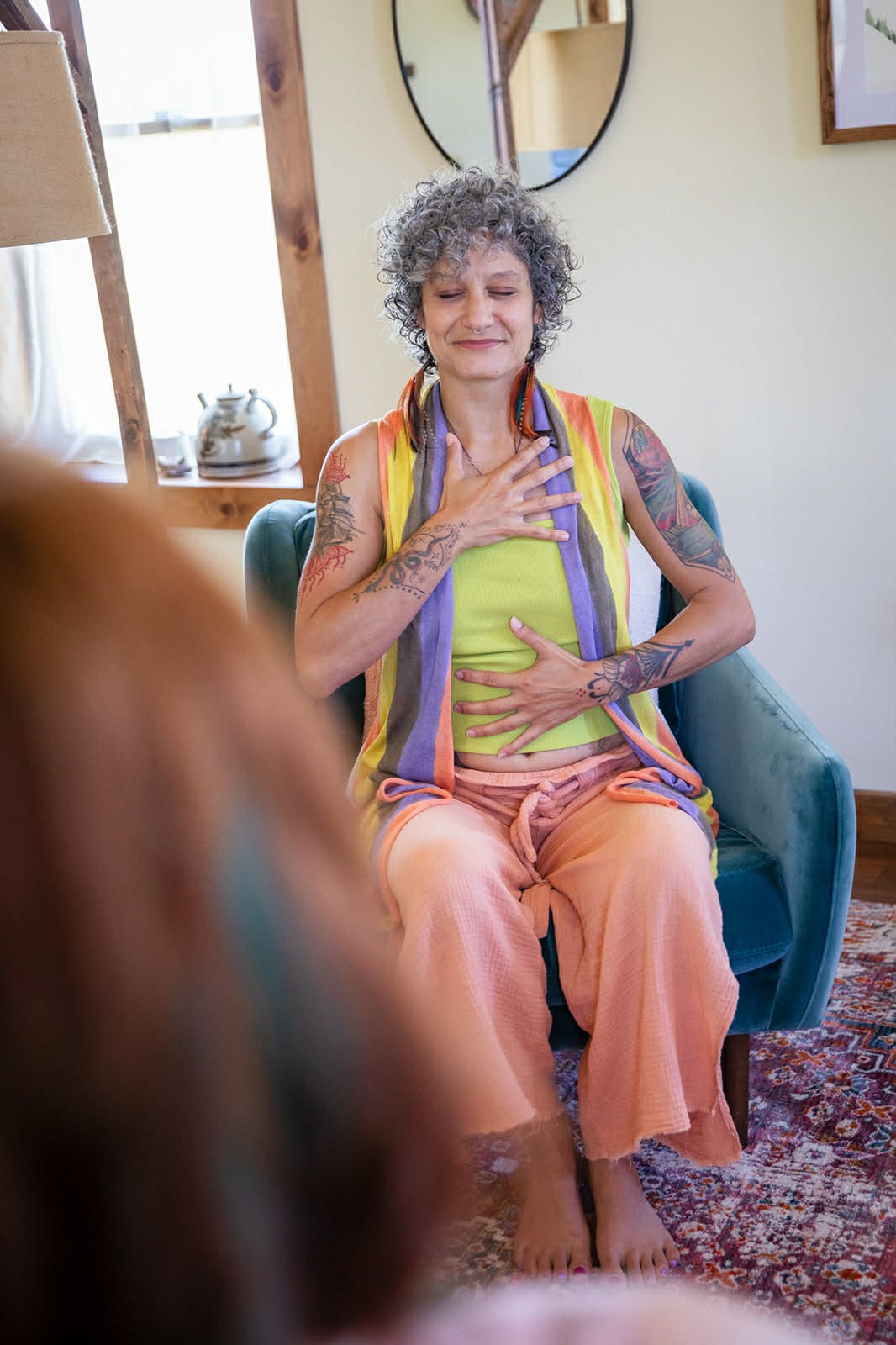 A woman sitting on a chair, smiling and holding a book, in a cozy, sunlit indoor space.