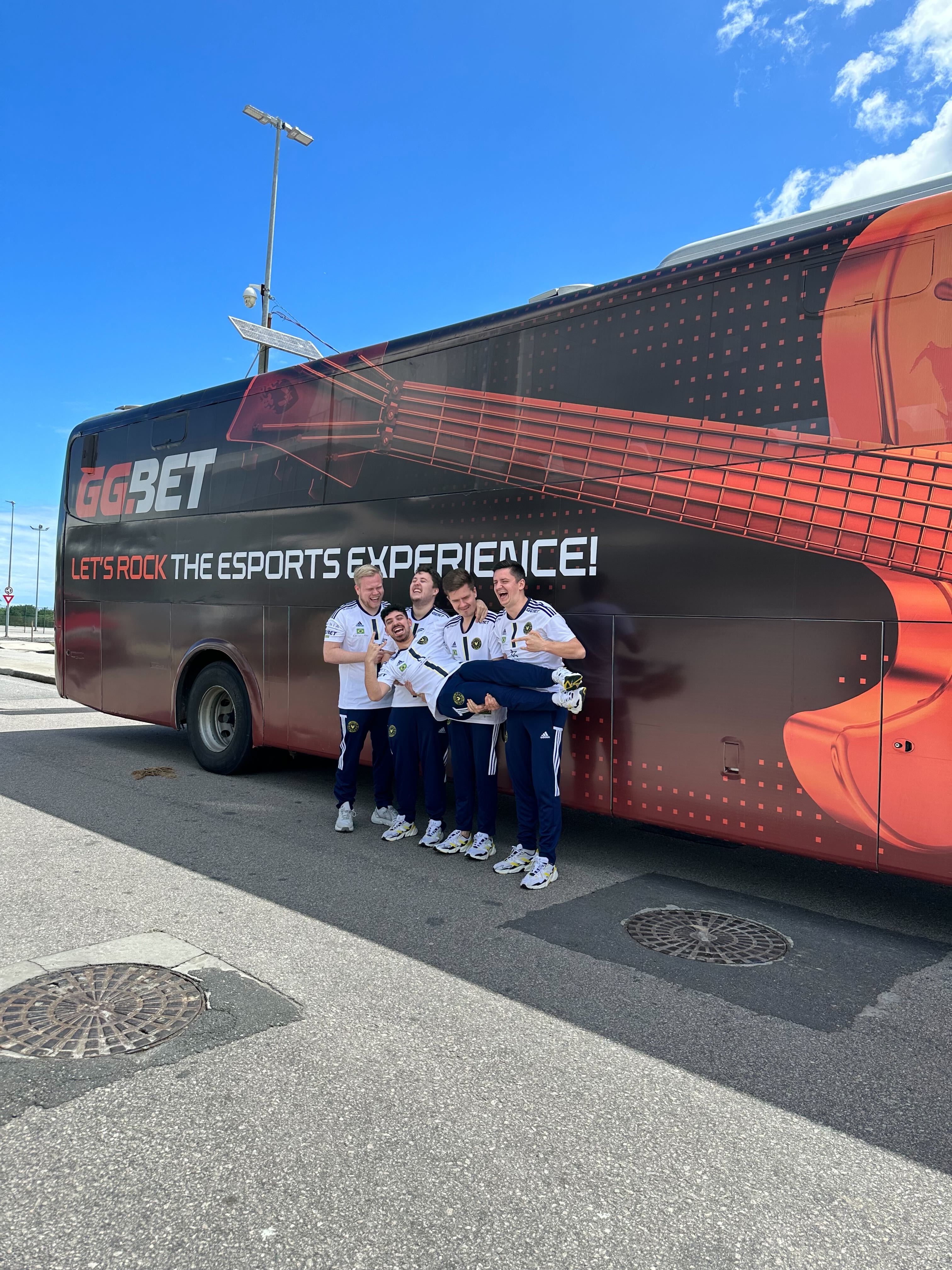 Visuel du projet Metality : l'équipe Metality devant leur bus de tournée rouge et noir. Sur le bus, un manche de guitare et le texte « Rockons l'expérience esports - GGBET » en rouge et blanc.