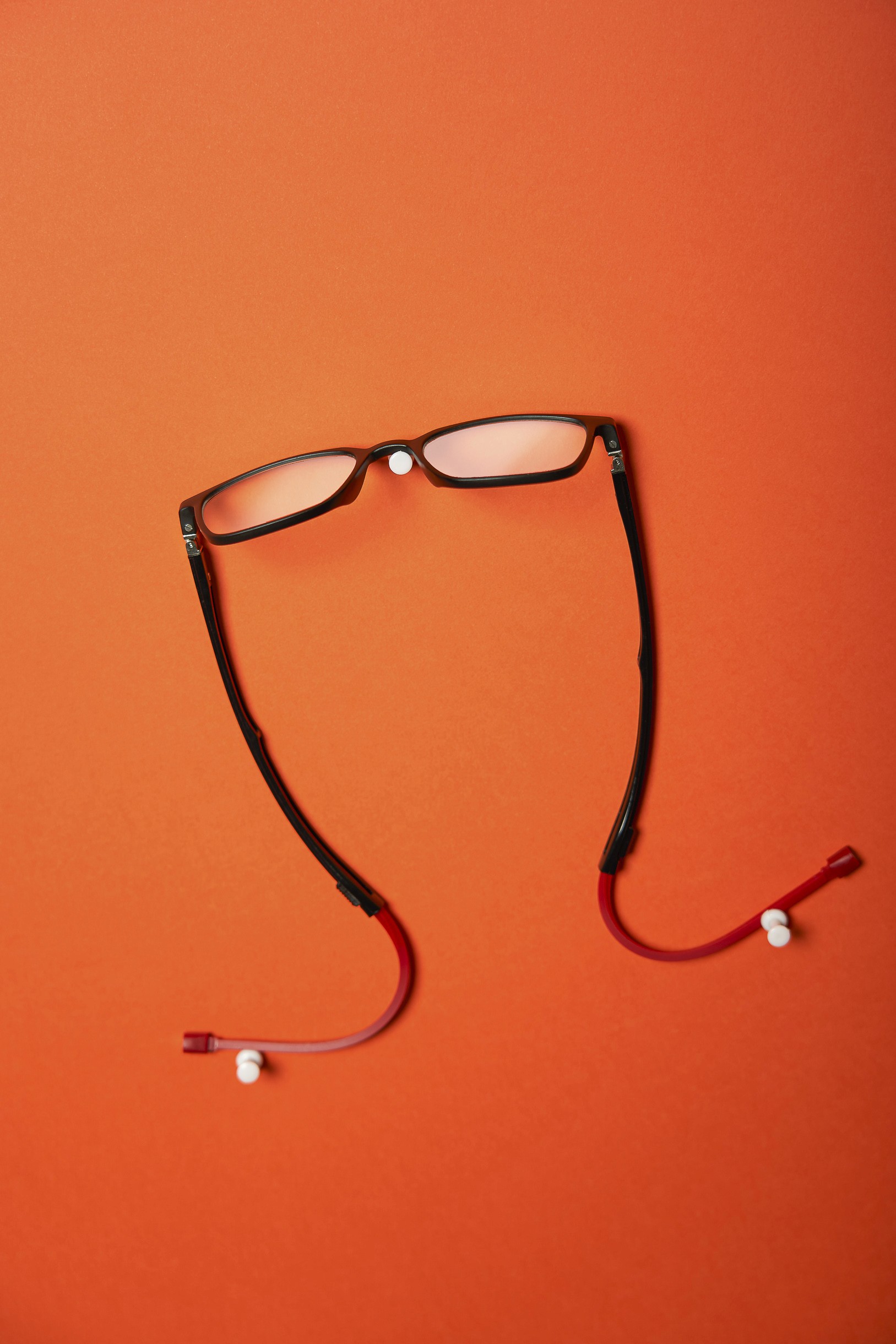 Photographie studio sur fond vif orange d'une paire de lunette de lecture noire accrochée au mur grâce à des punaises