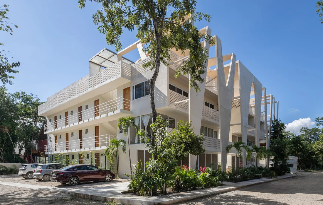 Edificio contemporáneo blanco de varios pisos con balcones, pilares en V y vegetación tropical, bajo cielo azul con autos estacionados
