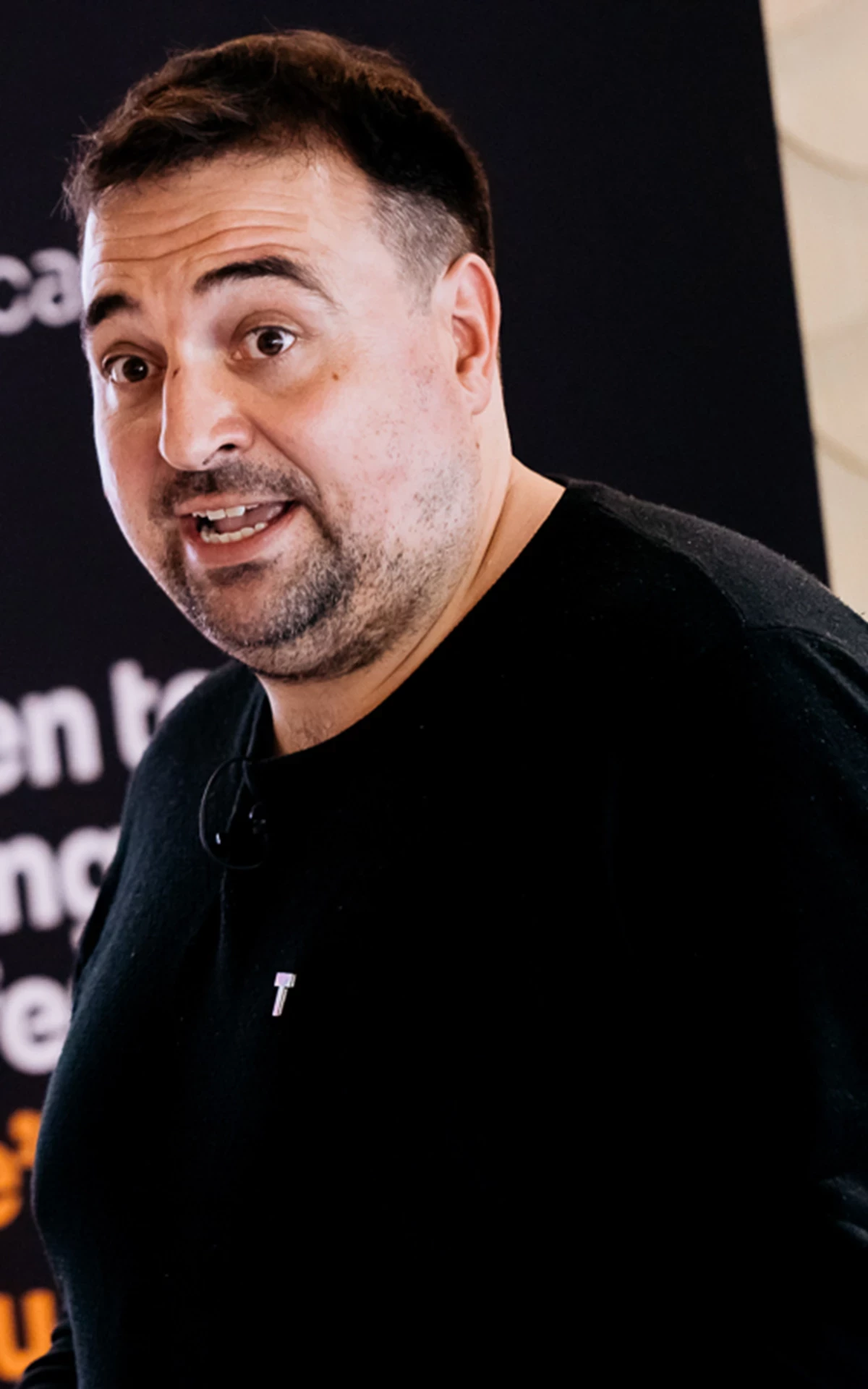 Person wearing a black long-sleeve shirt with a small ‘T’ logo, standing in front of a banner with text about doing tech differently.