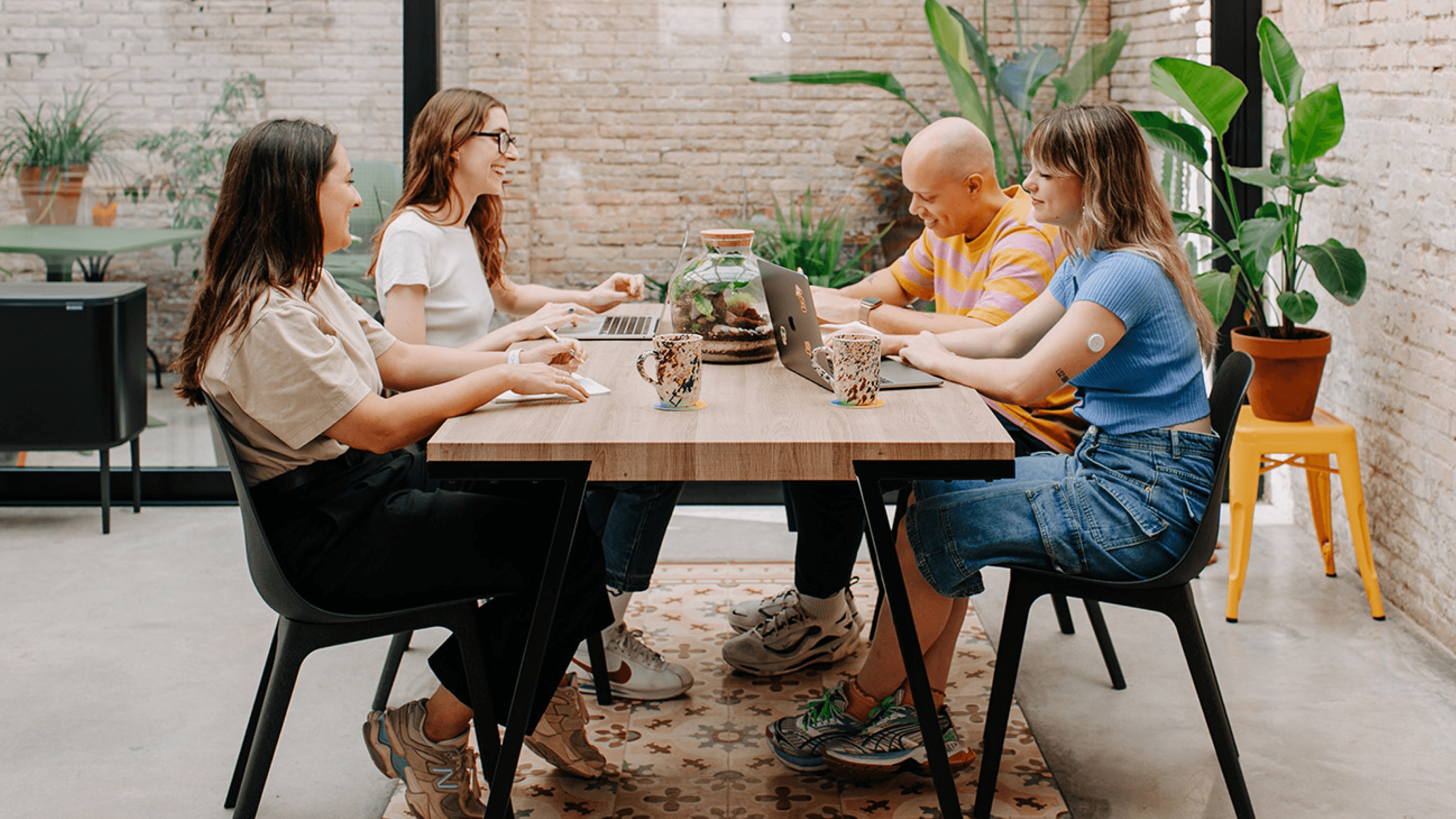 Four people sitting at a table working on their laptops while talking and smiling.