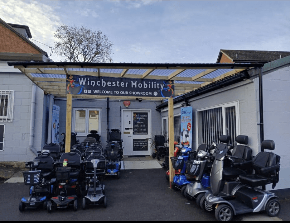 image shows a range of mobility scooters  lines up outside the front of the showroom. There are silver, blue and green scooters in a variety of sizes with bright starts on htem showing their prices and key information.