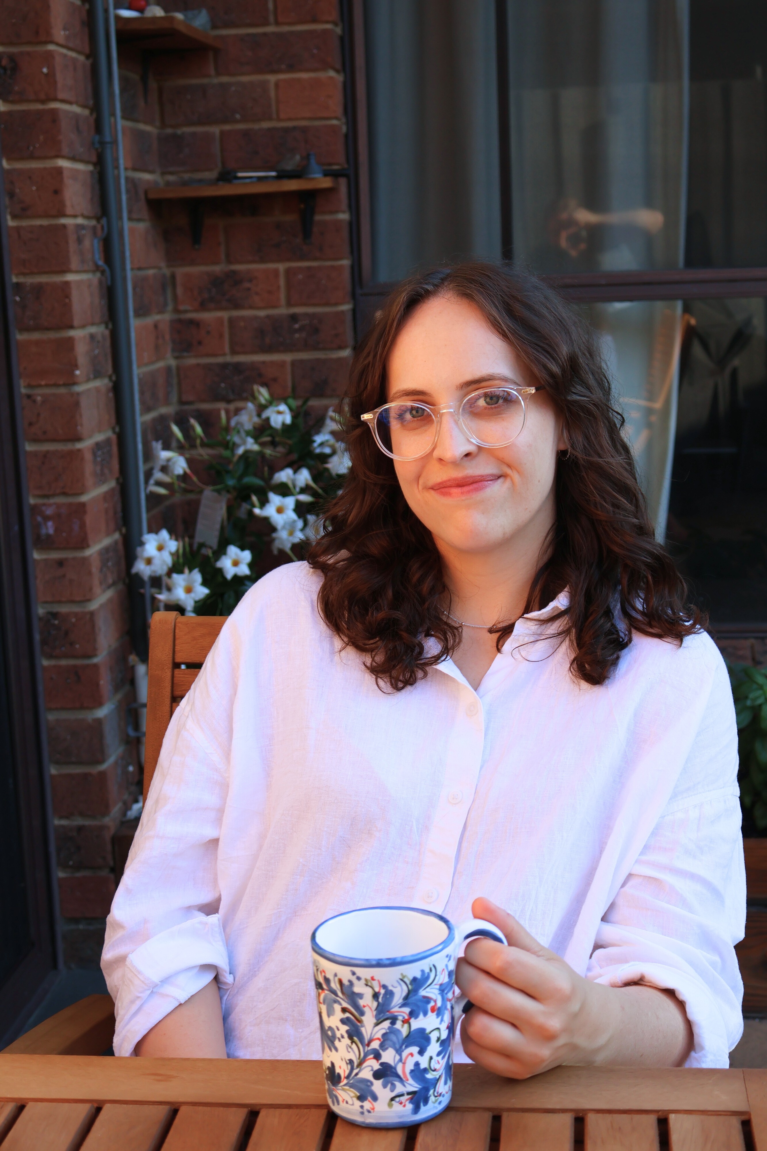 Woman in her late 20s with brown curly hair. She is wearing clear-rimmed glasses, a white shirt. She is sitting at a wooden table outside, with flowers in the background, nursing a blue and white mug of tea. She is smiling with her mouth closed, looking at the camera.