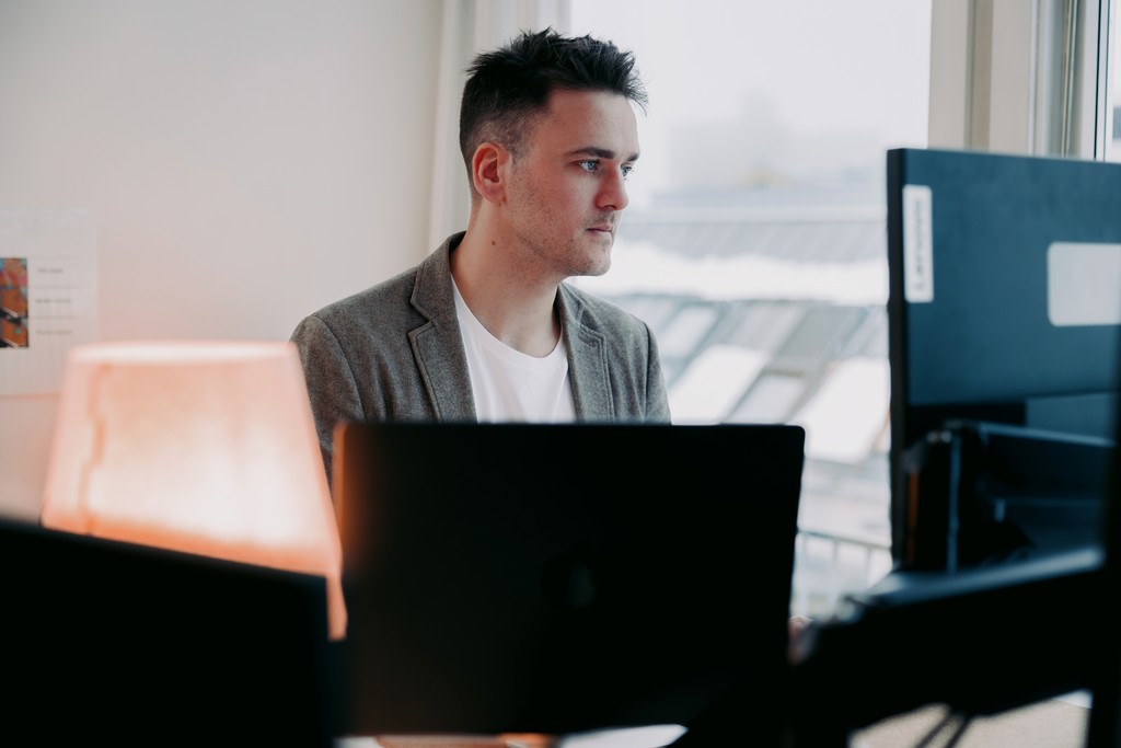 A person in a grey blazer and white shirt focuses intently on a computer screen in a modern office setting with muted lighting and a warm desk lamp nearby, suggesting a professional work environment.