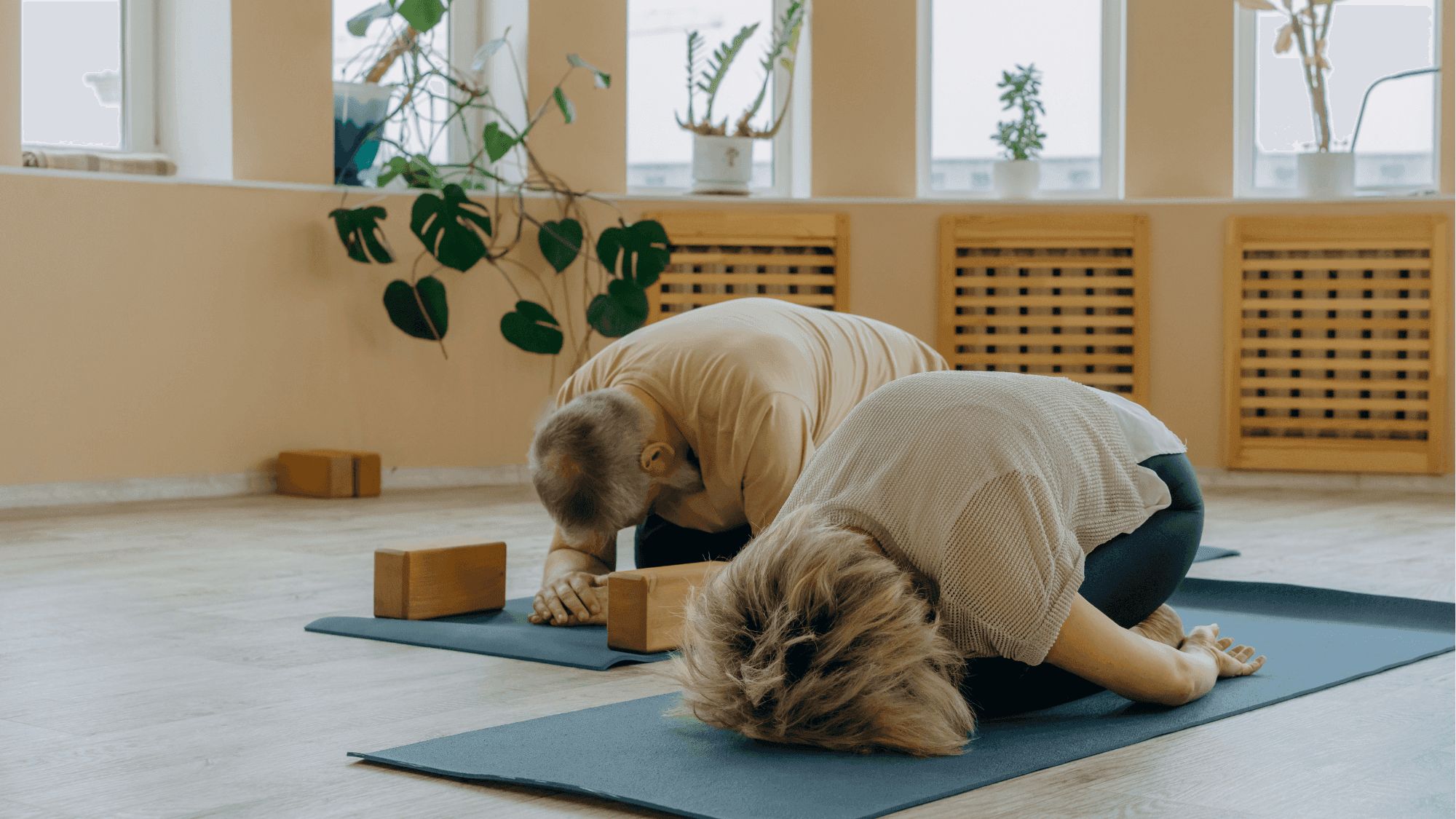 Two people practicing Child's Pose (Balasana) on blue yoga mats in a serene studio with monstera plants and wooden yoga blocks, demonstrating proper restorative yoga alignment"