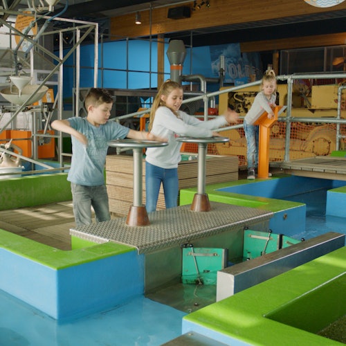 三个孩子在室内科学或活动中心参与互动展览，这些展览涉及水和机械元素。