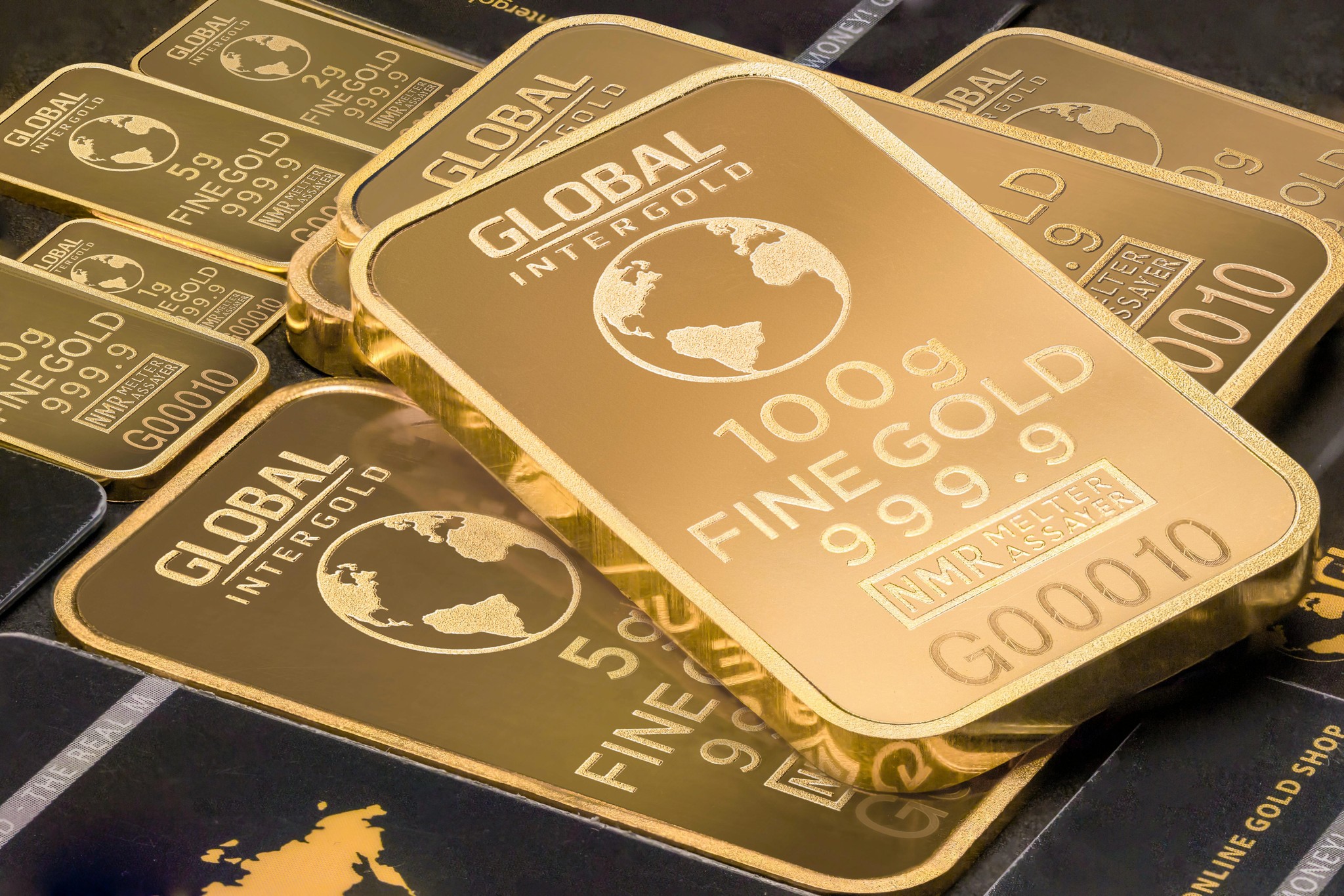 A close-up shot of several shiny, minted gold bars stacked overlapping each other. The largest bar in the foreground is engraved with "GLOBAL INTERGOLD" and a world globe logo, followed by "100g FINE GOLD 999.9", an "NMR MELTER ASSAYER" stamp, and serial number "G00010". Other smaller bars in the background are labeled with weights like 1g, 2g, and 5g. The bars feature a polished, reflective finish with frosted lettering and sit atop textured black surfaces.