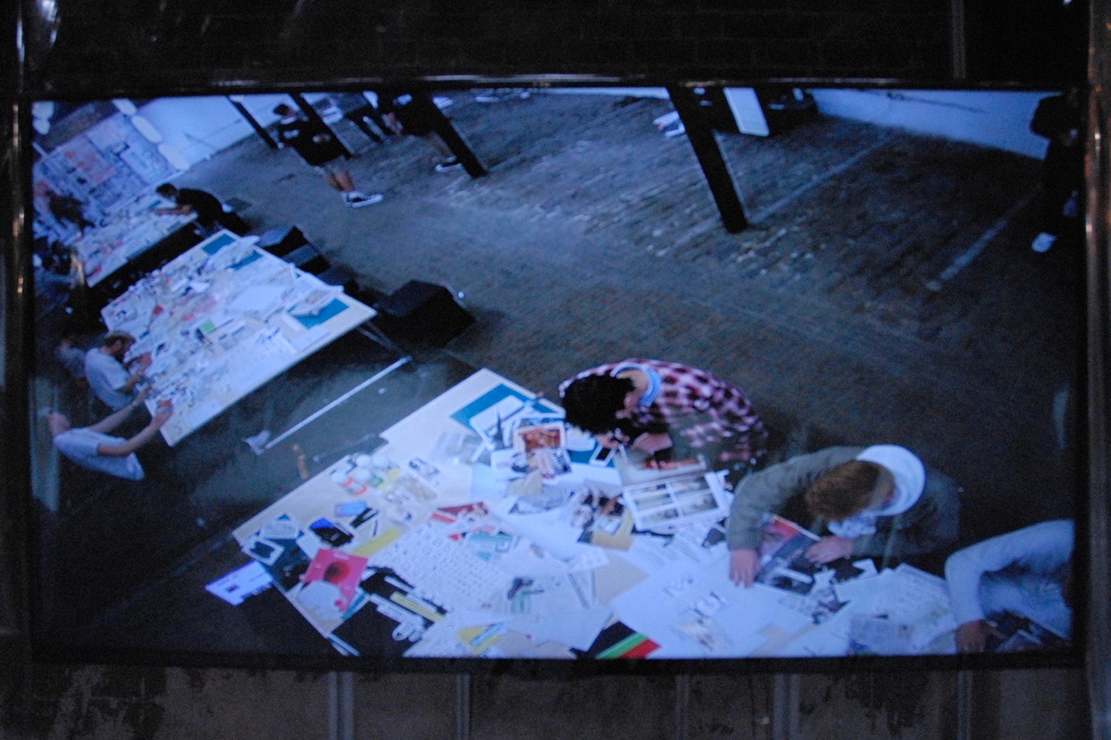 Aerial view of a creative workshop with several people engaged in art projects at tables covered with papers, sketches, and art supplies in a spacious, industrial-style room.