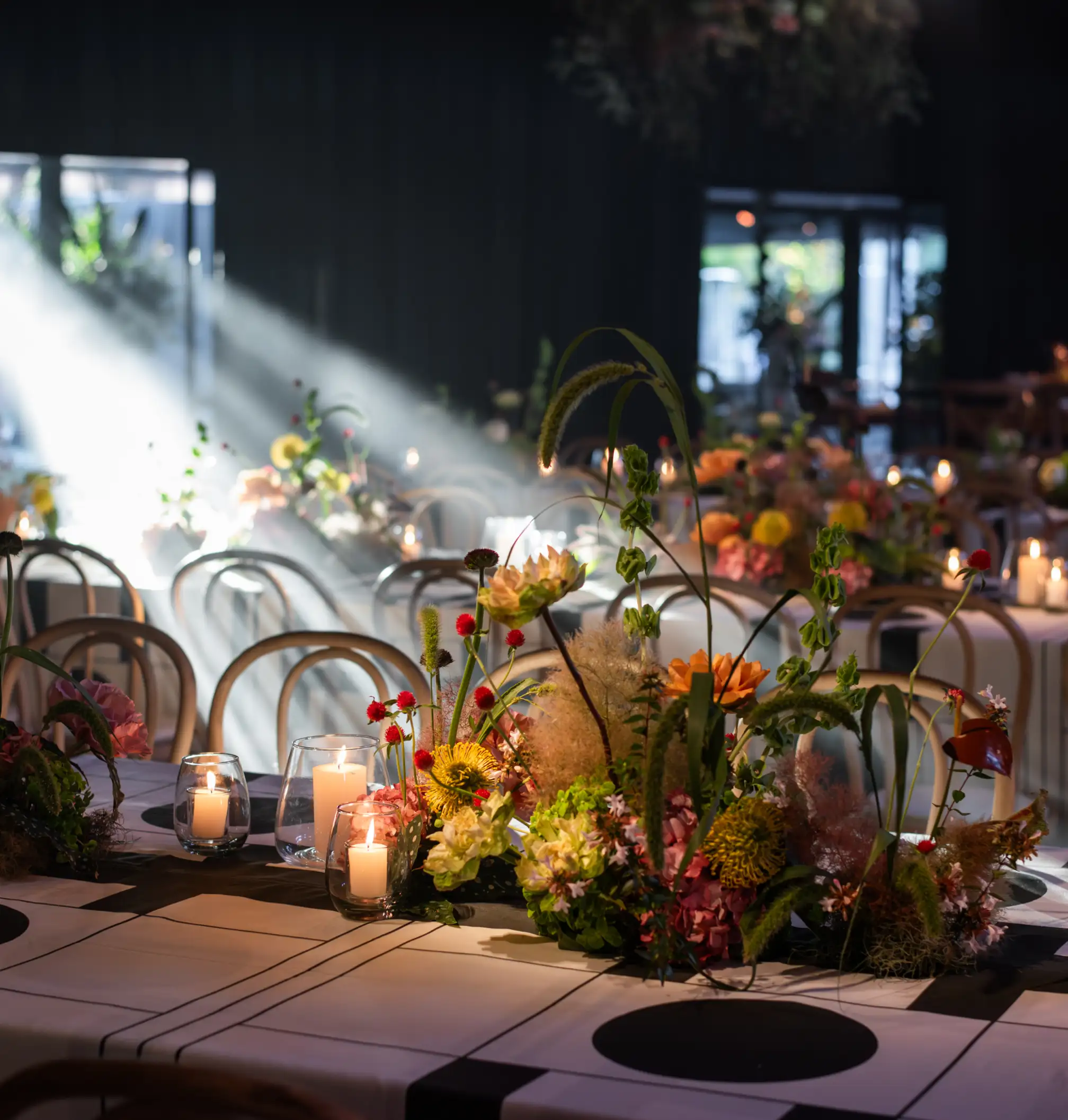 Mesa de evento iluminada por un haz de luz, con centros florales coloridos y velas encendidas sobre manteles gráficos en blanco y negro para ambientación moderna y teatral.