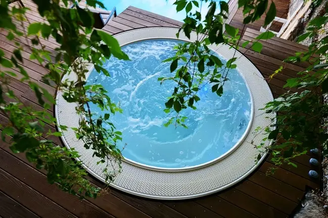 Runder Whirlpool auf der Holzterrasse von oben, umrahmt von grünen Blättern.