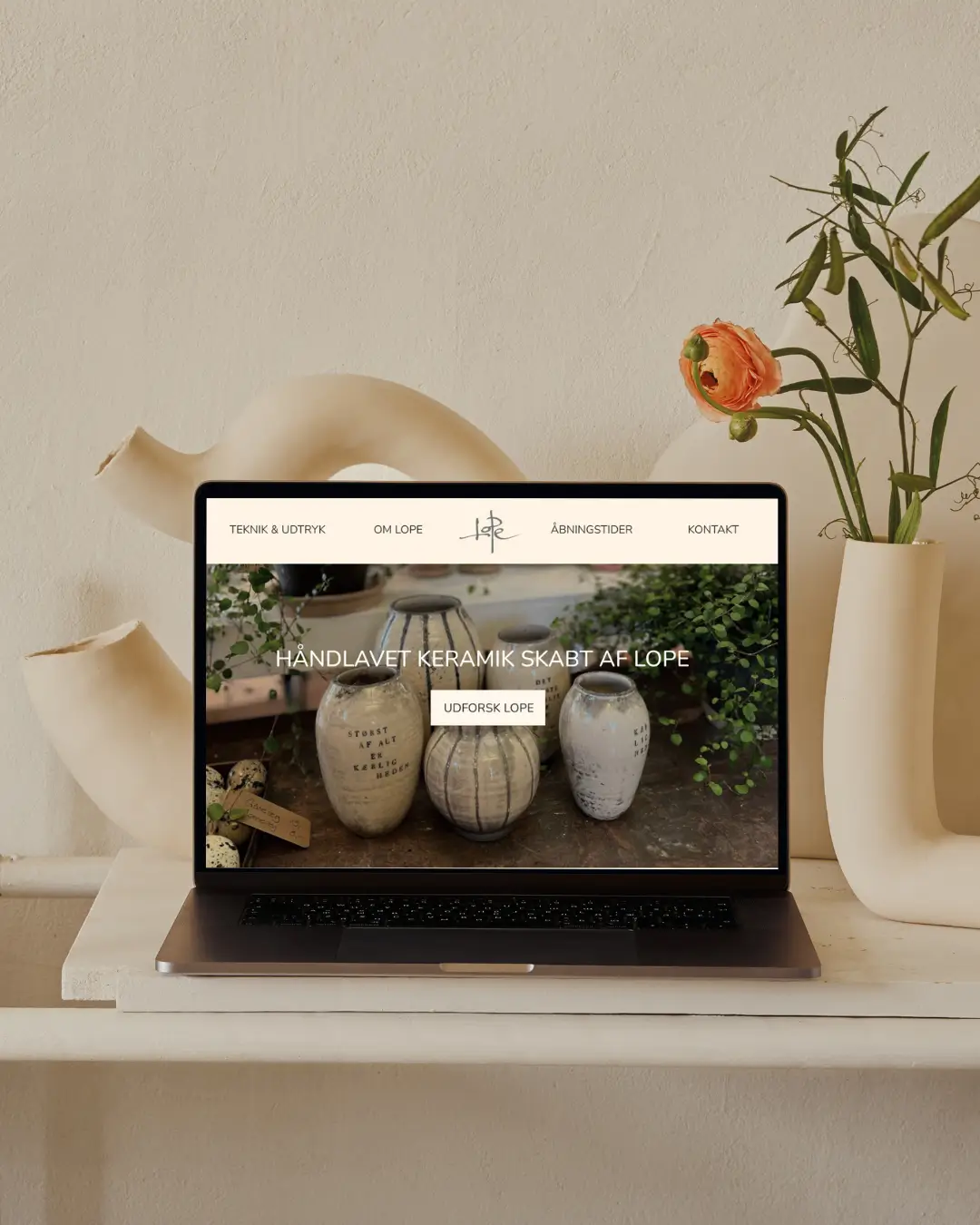 Computer der viser håndlavet keramik hjemmeside, omgivet af vaser og blomster.
