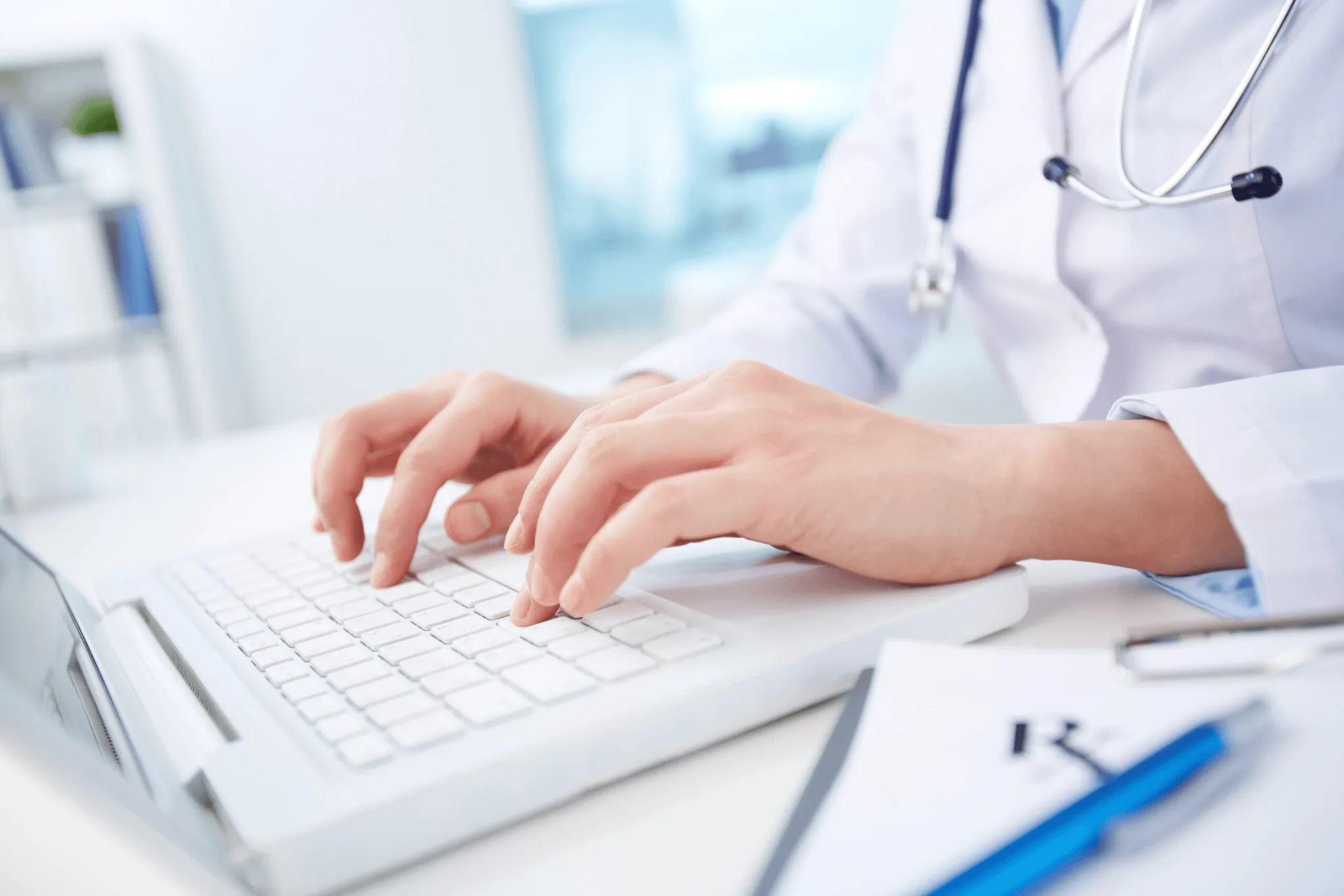 Hands of a physician wearing a white coat and stethoscope around the neck, typing in a laptop, pen and checklist board beside.