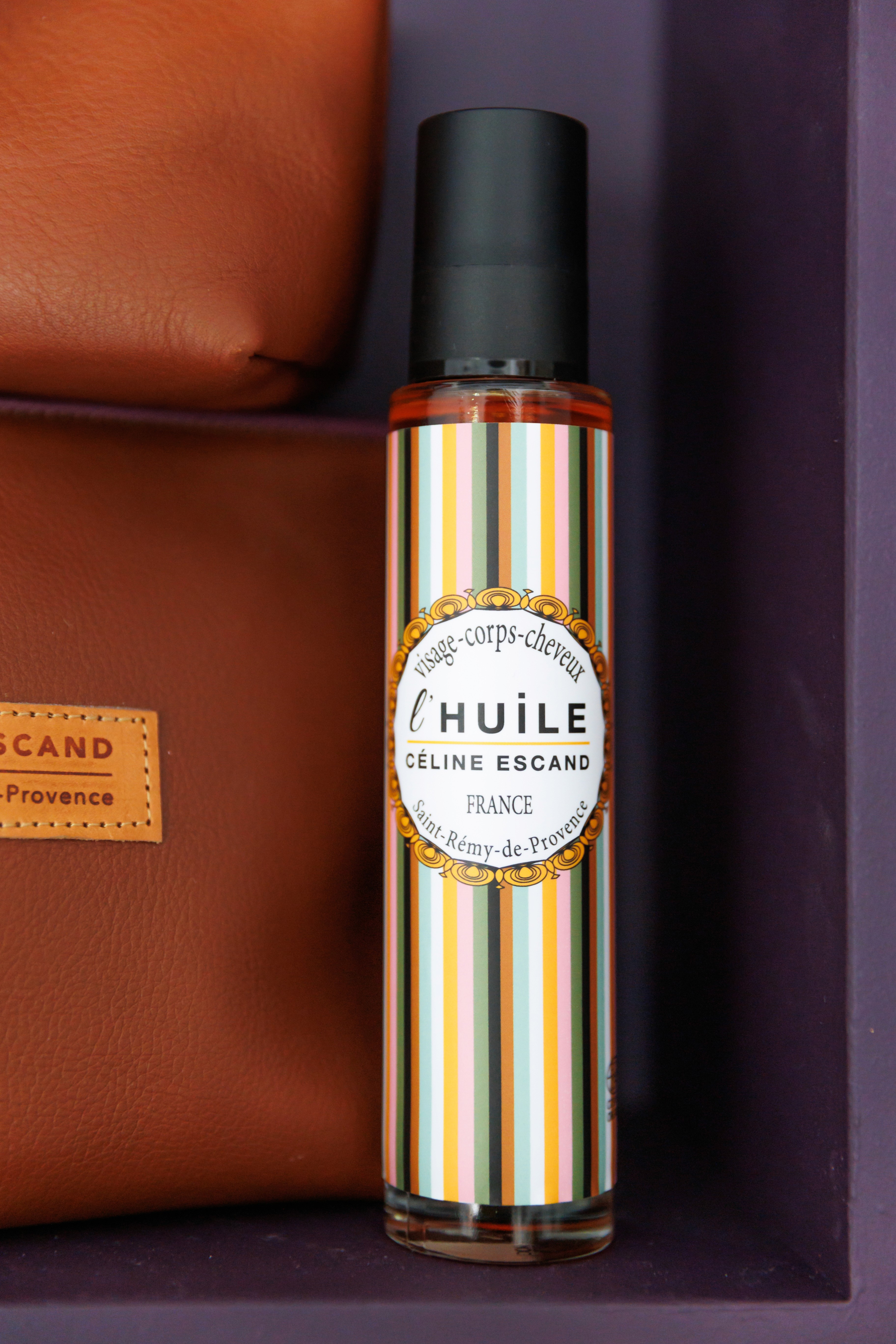 Flacon d’huile LBI Céline Escand posé à côté d’une pochette en cuir marron, design à rayures colorées, photographie produit élégante et soignée.