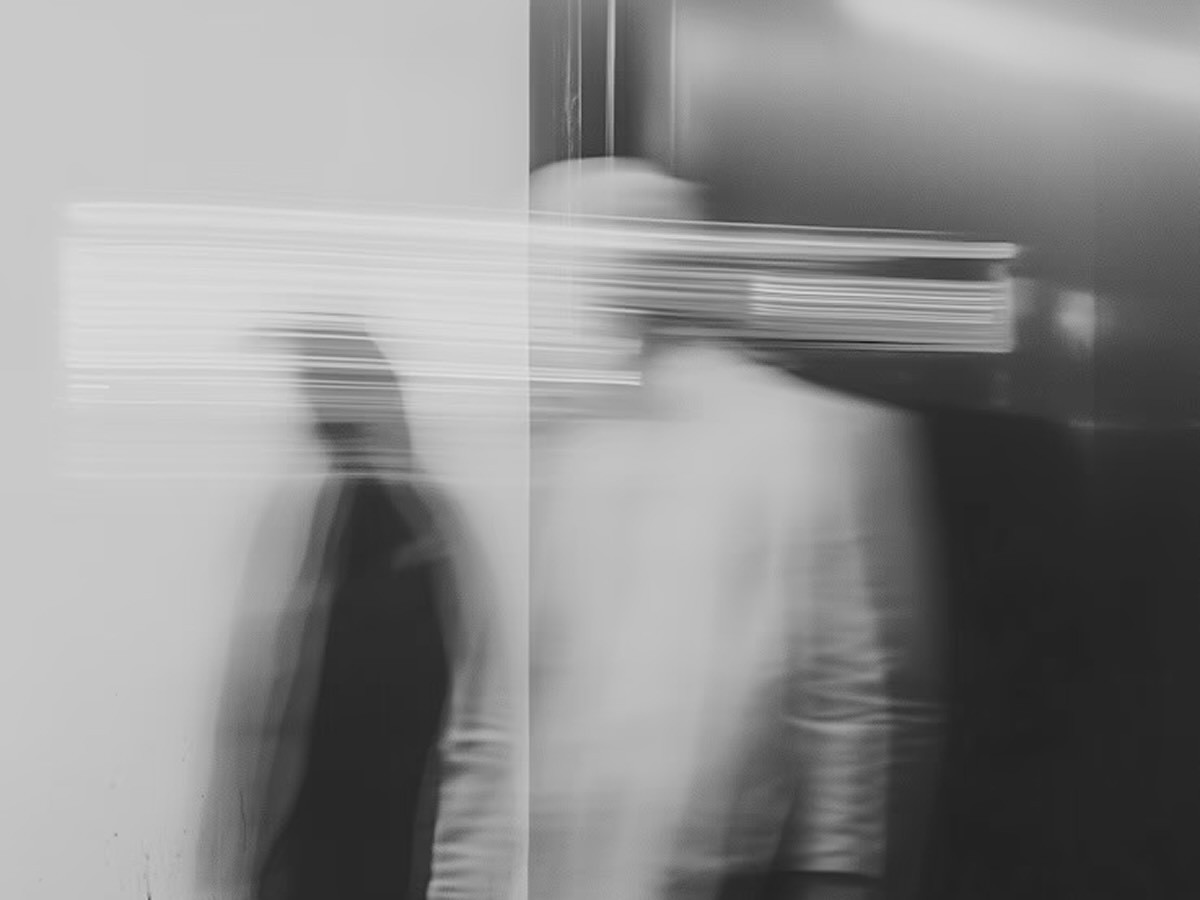 Black-and-white motion-blurred image of two indistinct figures standing in a stark interior beside a reflective glass surface.