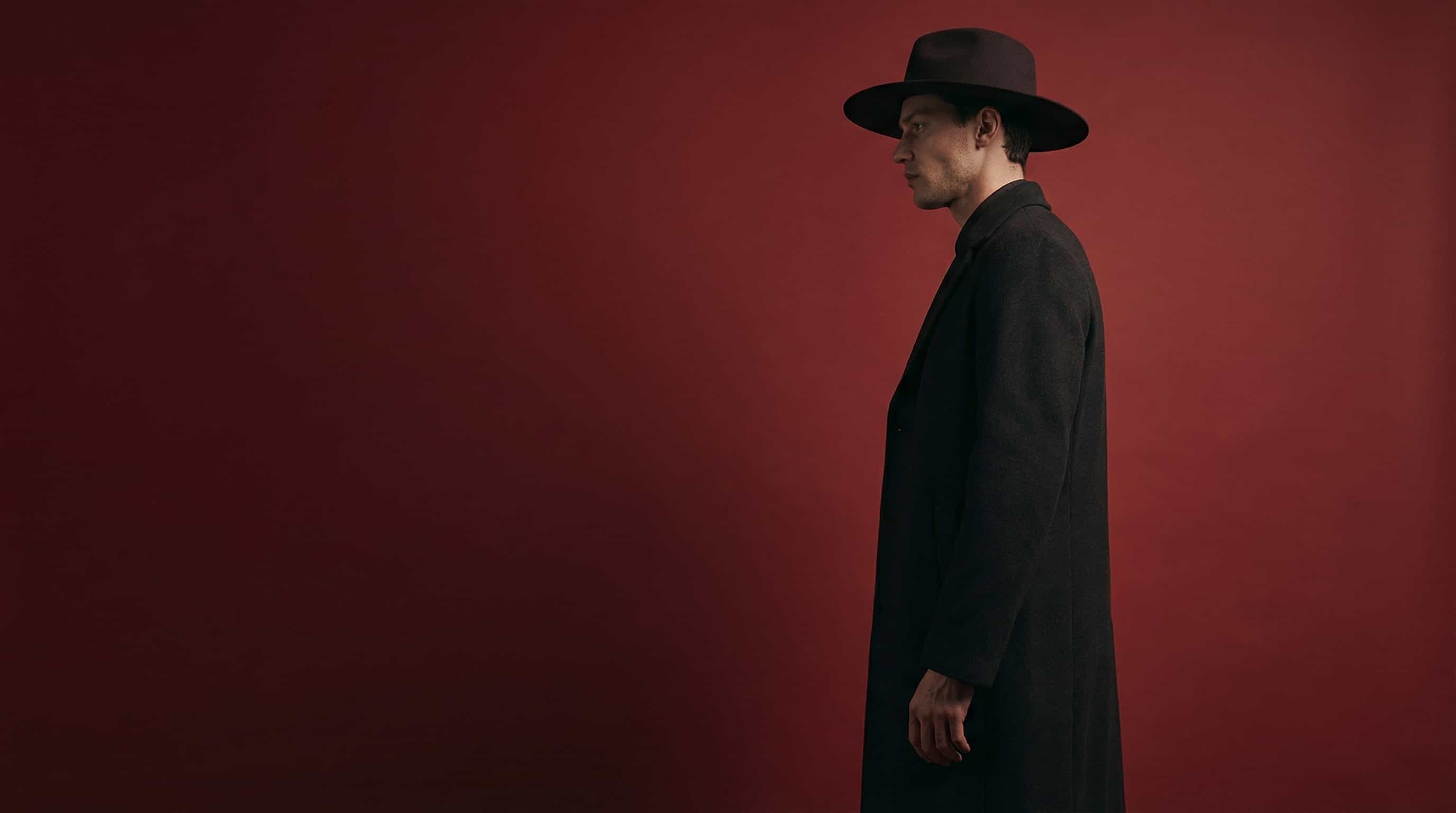 Man in a dark coat and hat standing against a red studio backdrop.