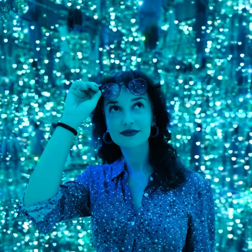 A woman in a sparkly shirt stands against a backdrop of blue, shimmering lights, adjusting her sunglasses while looking upward.