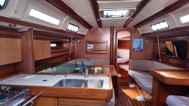 Dufour 40 Performance sailing yacht main saloon interior looking forward, showing the stainless galley double sink with chrome tap, warm cherry wood cabinetry, teak-and-holly sole, forward cabin door with arched trim, port settee with grey cushions, overhead deck hatch, and multiple hull portlights