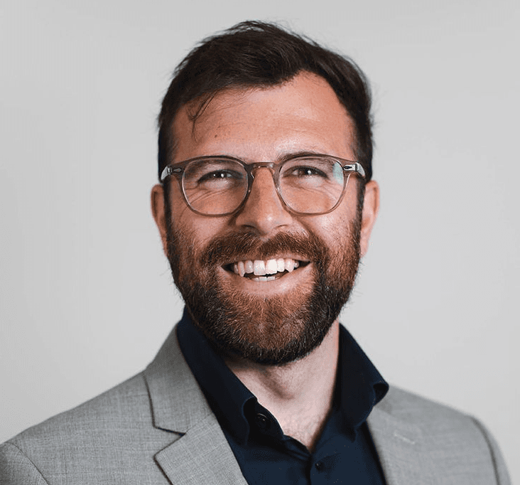 Person wearing a blue puffer jacket and red backpack standing in front of a light blue building on a city street.