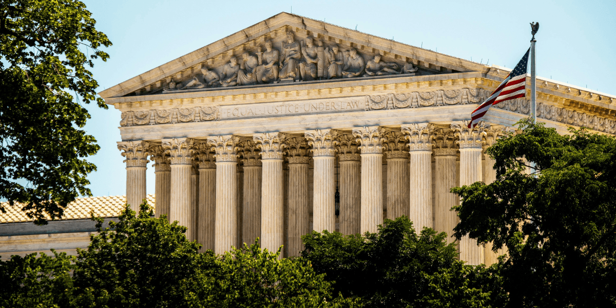 The image shows the U.S. Supreme Court building, with tall columns and detailed carvings. An American flag waves in the foreground under a clear sky.