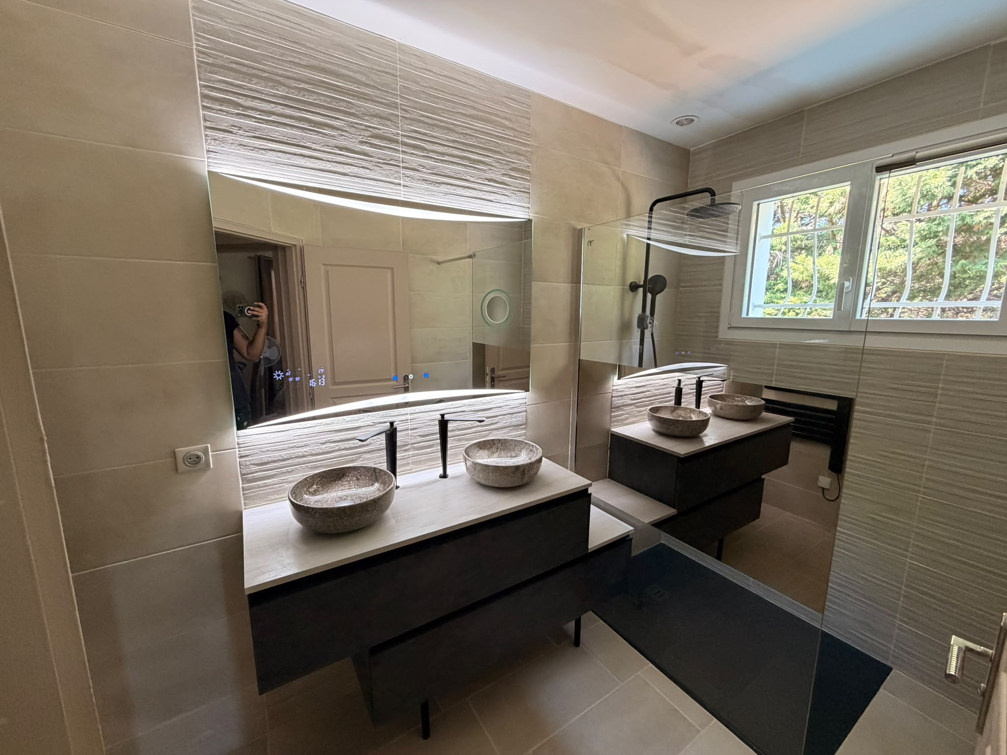 Salle de bain rénovée avec double vasque en pierre, meuble sombre, miroir rétroéclairé et douche vitrée à robinetterie noire.