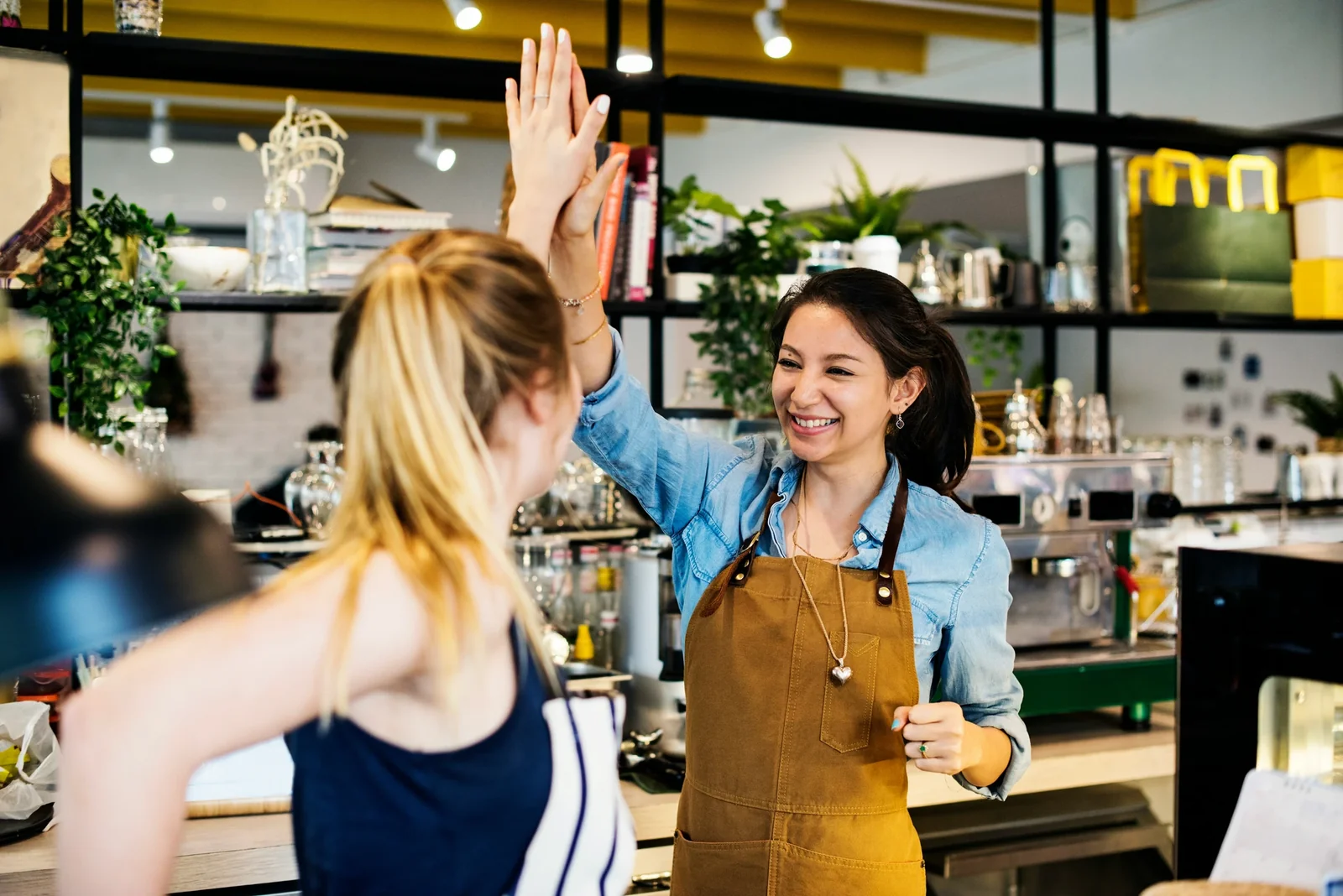 Two women high-fiving at a coffee shop counter, smiling and celebrating together in a bright, modern café setting.