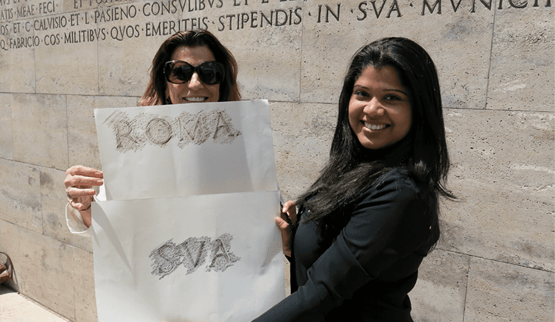 A photo of two women standing near a letter carved stone wall, while holding pieces of paper with text ROMA SVA. The text has ben transferred from the wall engraving to the paper.