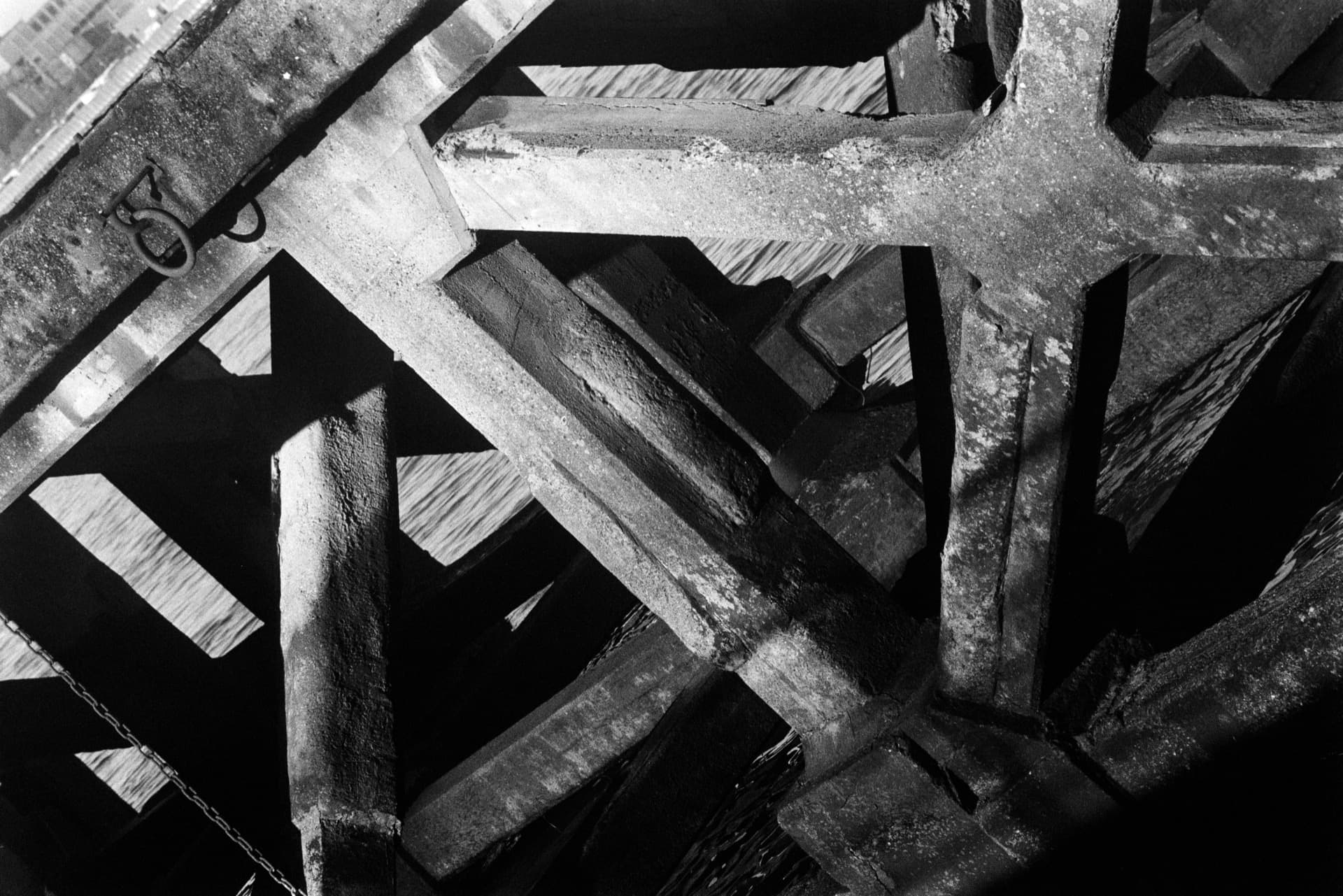 Angular concrete beams crossing at oblique angles beneath dock structure, creating diagonal composition of weathered surfaces and deep shadows